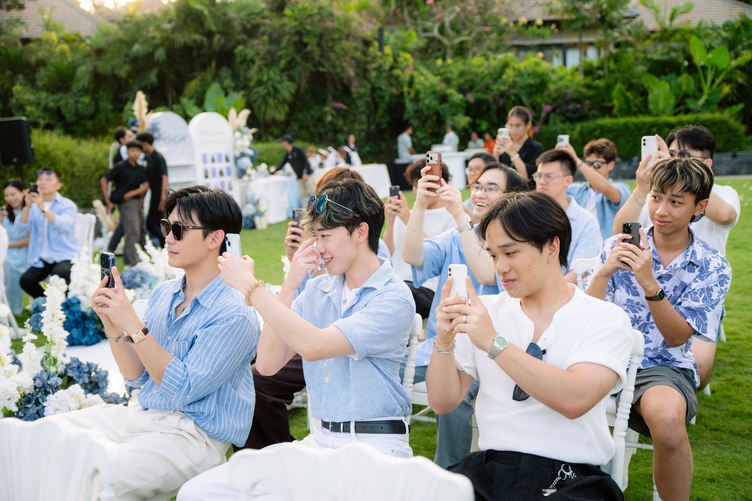 Wedding guests in light blue shirts sit on white chairs on a sunny lawn, holding up phones to film the outdoor ceremony among flowers.