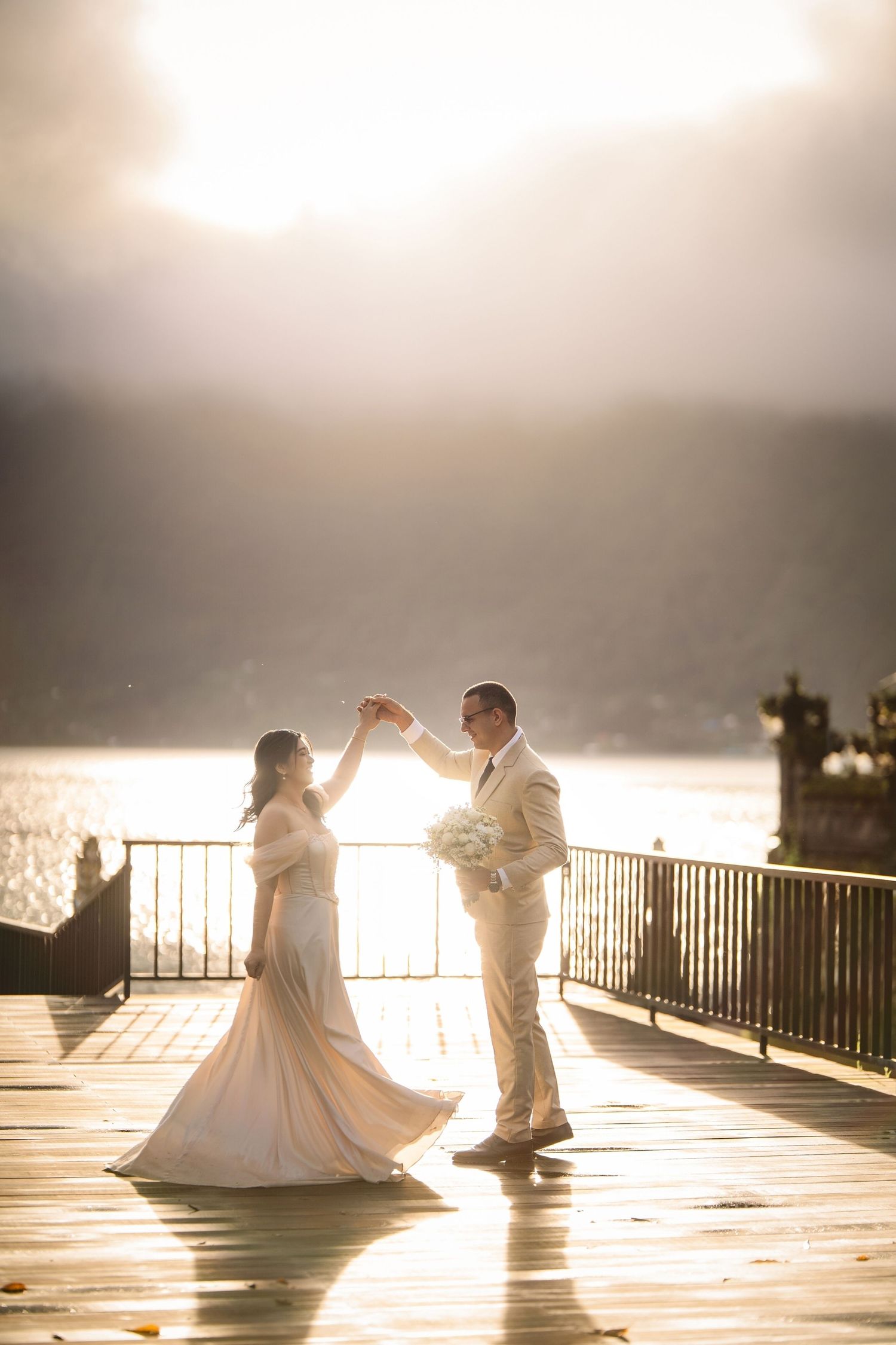 Bride and groom dancing on a sunlit waterfront deck with mountains and golden light in the background.