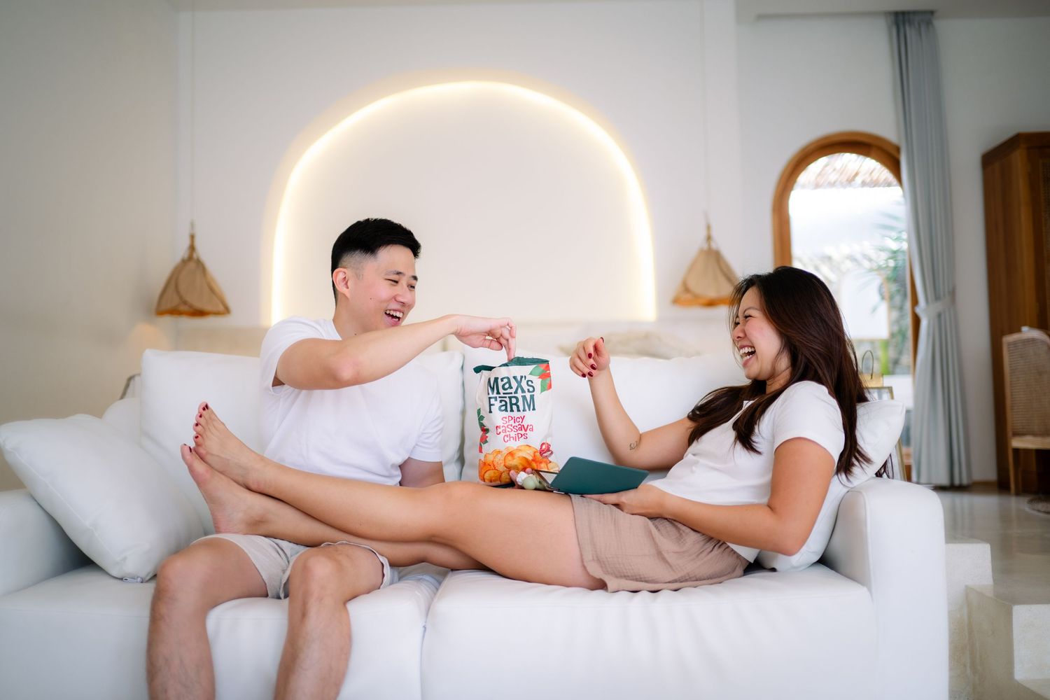 Couple sitting on white sofa sharing snacks and laughing together in a bright modern living room.