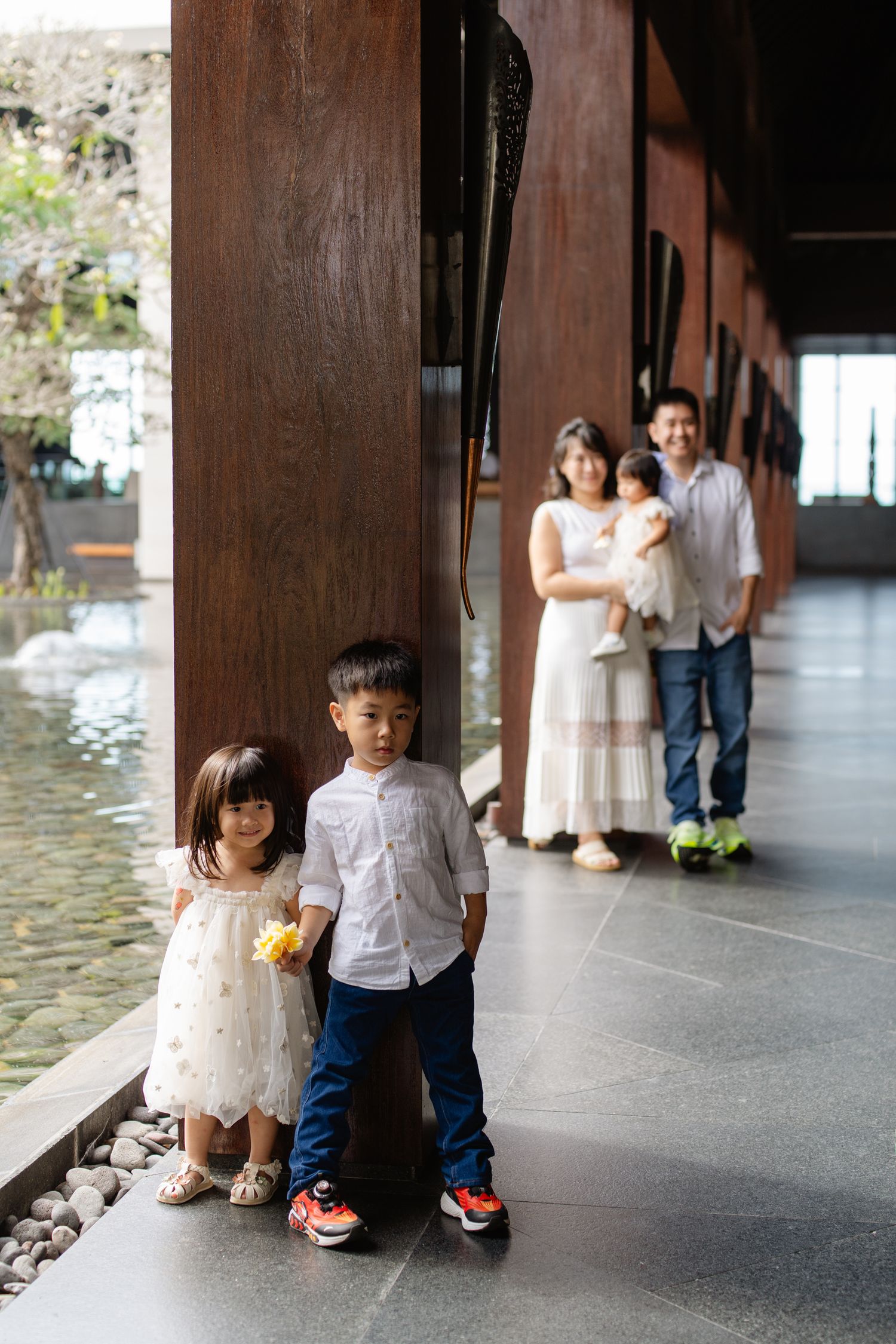 Asian family posing in modern corridor, two children in foreground, parents holding baby in background.