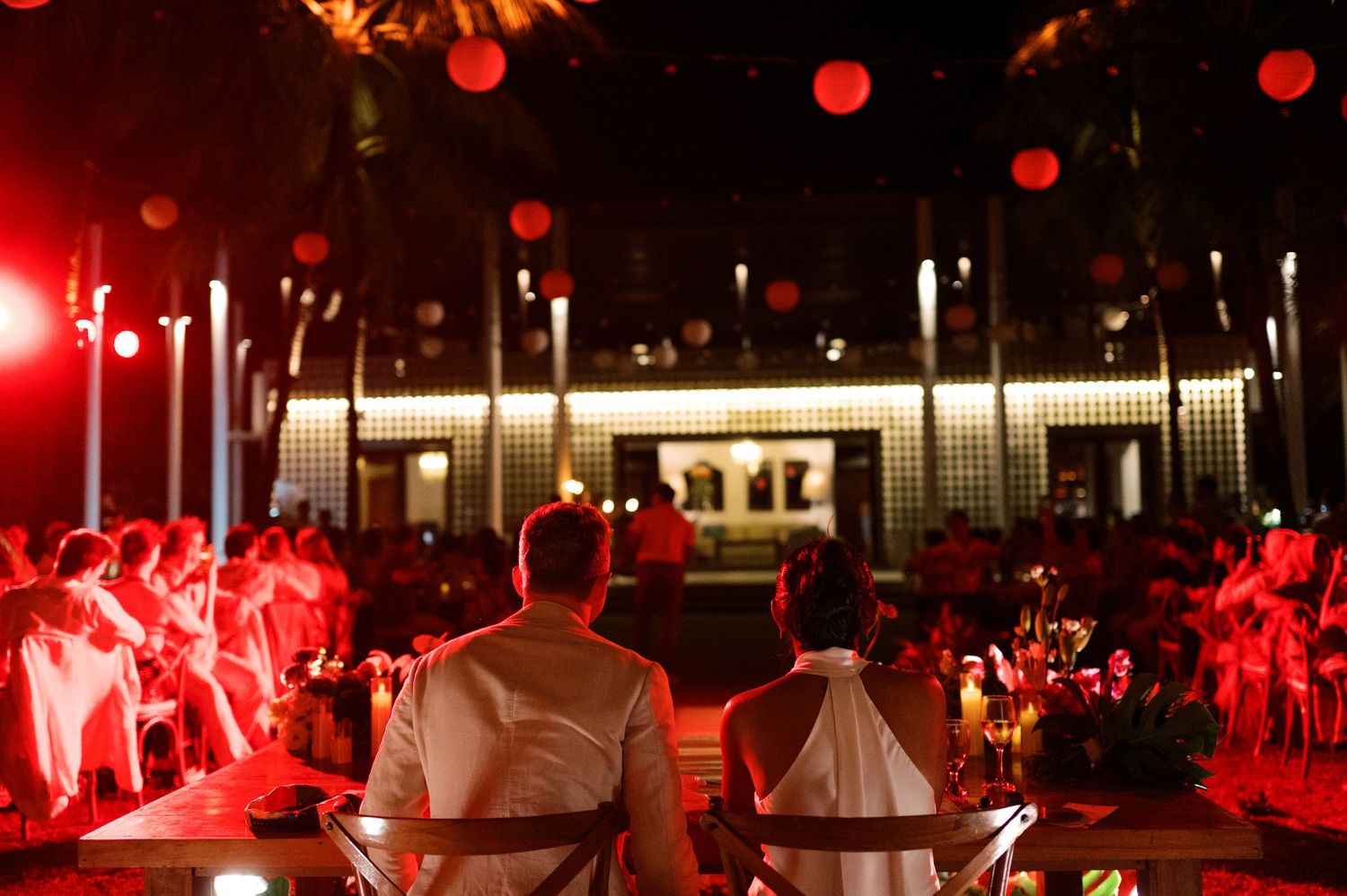 Couple seated at wedding reception table with red lanterns and warm lighting at outdoor evening event.