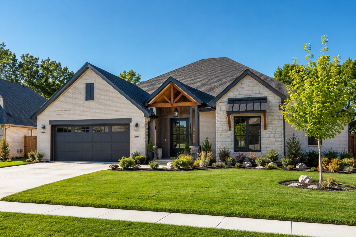 Modern craftsman-style home with stone facade, dark garage doors, wood accents, and lush green landscaping under a clear blue sky.
