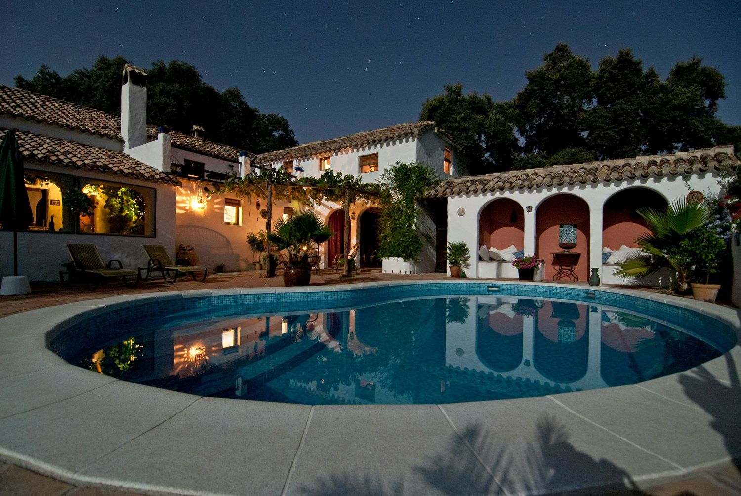 Elegant Spanish villa with oval swimming pool illuminated at dusk, featuring arched colonnades and lush tropical landscaping.