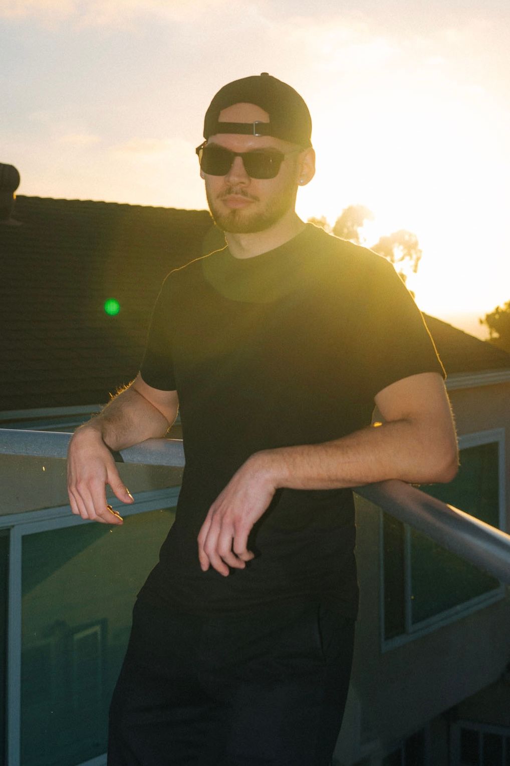 Young man in black outfit and sunglasses leaning on railing during golden sunset.
