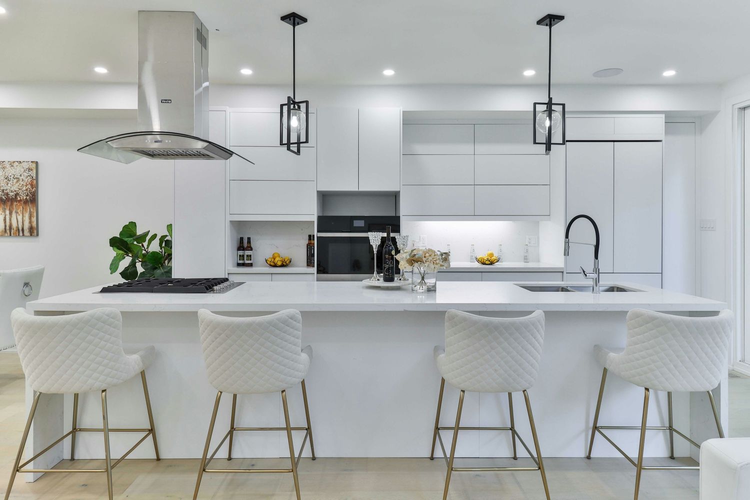 Modern white kitchen with large island, quartz countertops, gold bar stools, pendant lights, and stainless steel range hood.