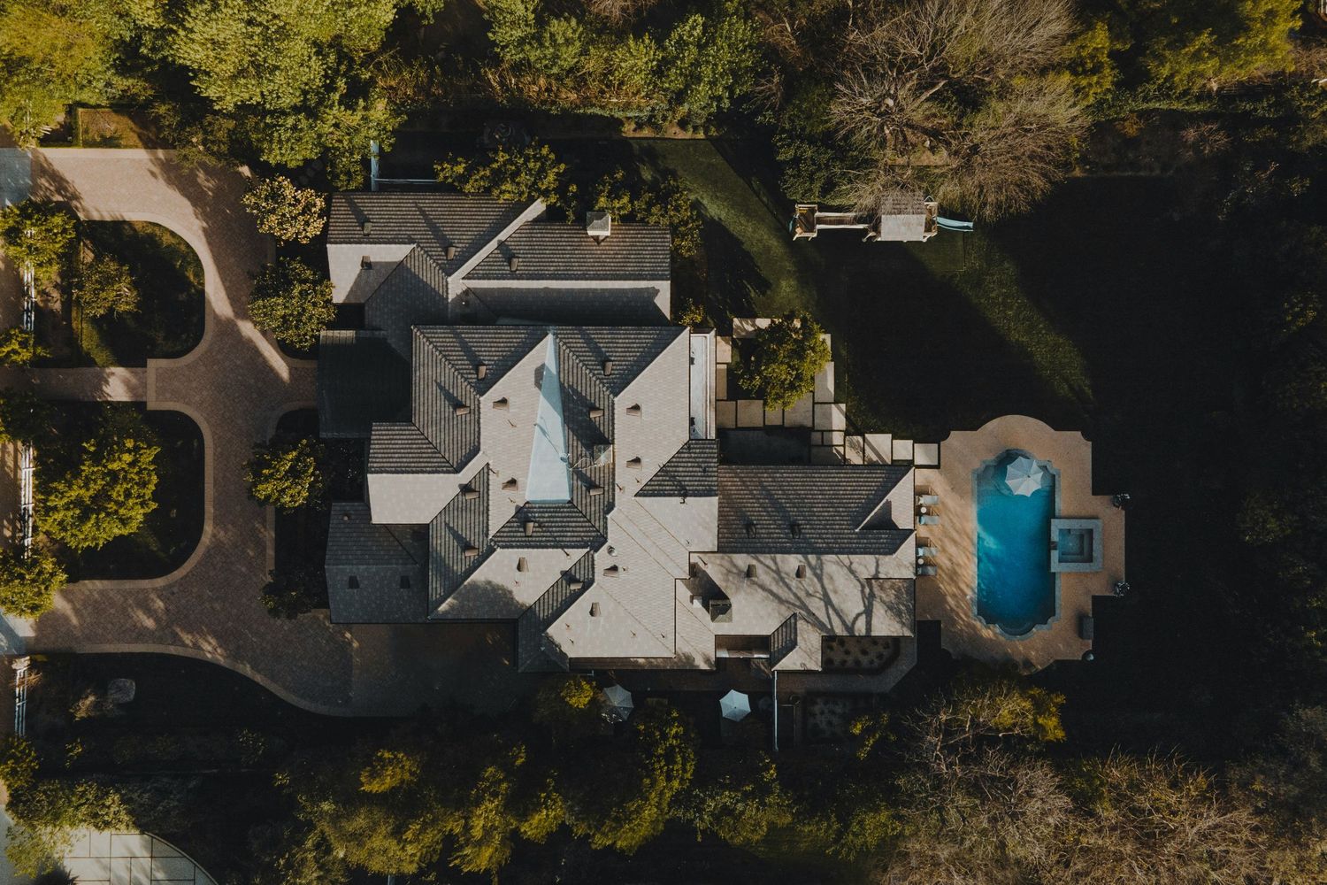 Aerial drone view of a large luxury estate with a gray roof, swimming pool, and lush surrounding trees.