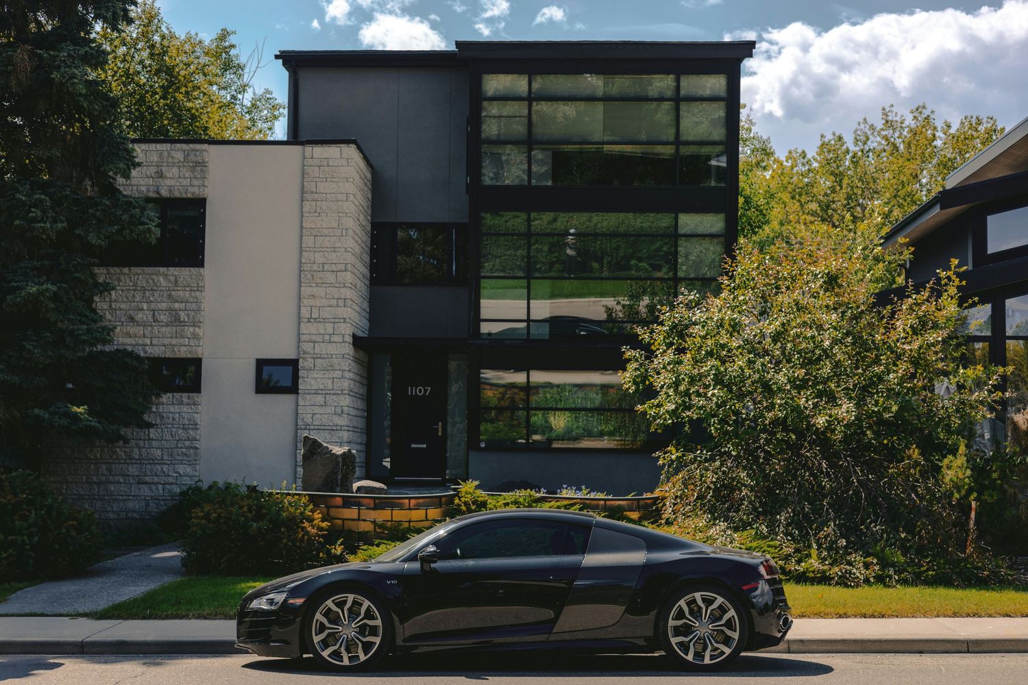 Black sports car parked in front of a modern two-story home with large windows and lush green trees.