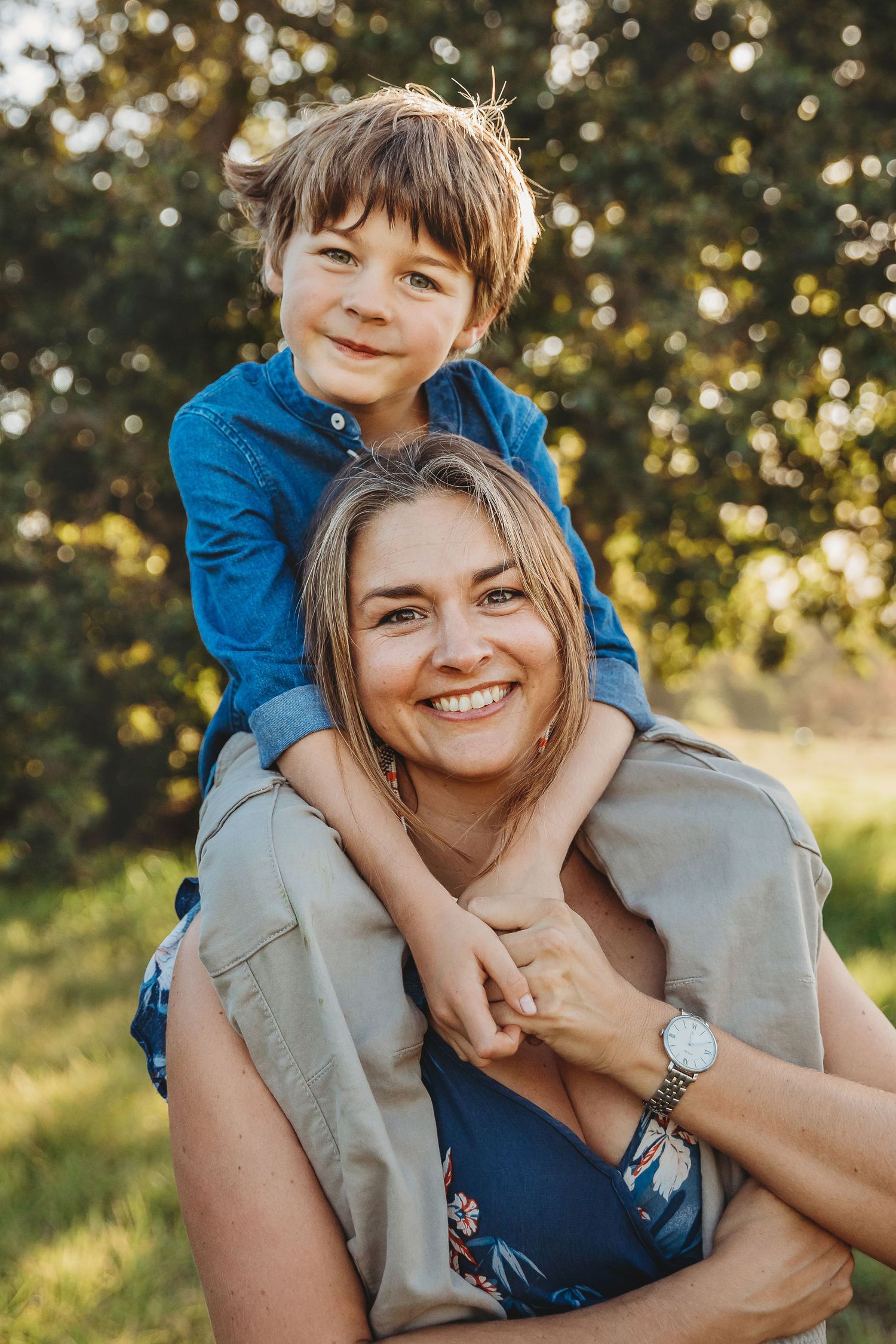 A smiling woman carries a happy young boy on her shoulders outdoors in a sunny green park setting.