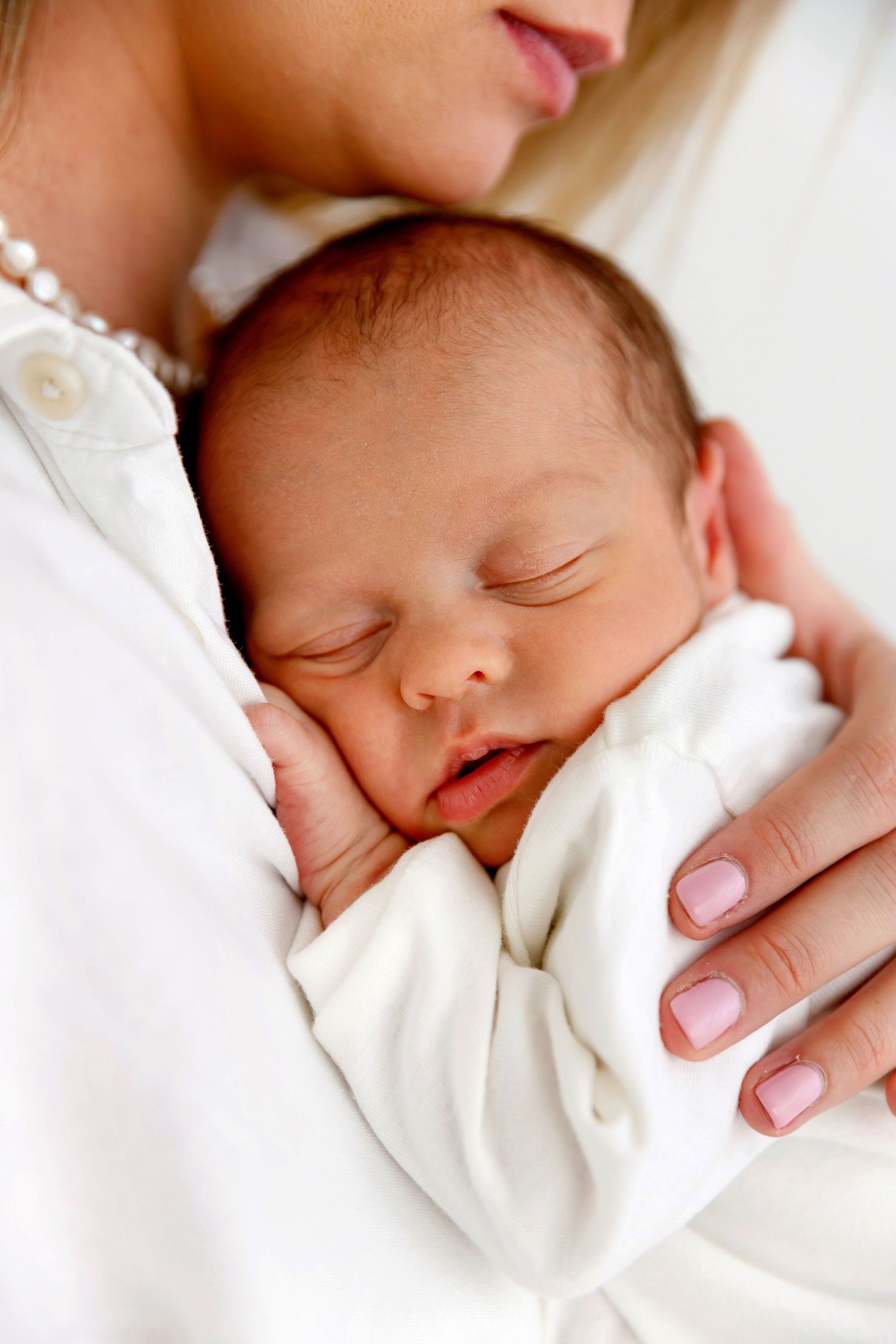 A mother in white gently cradles her sleeping newborn baby dressed in white, in a tender and peaceful moment.