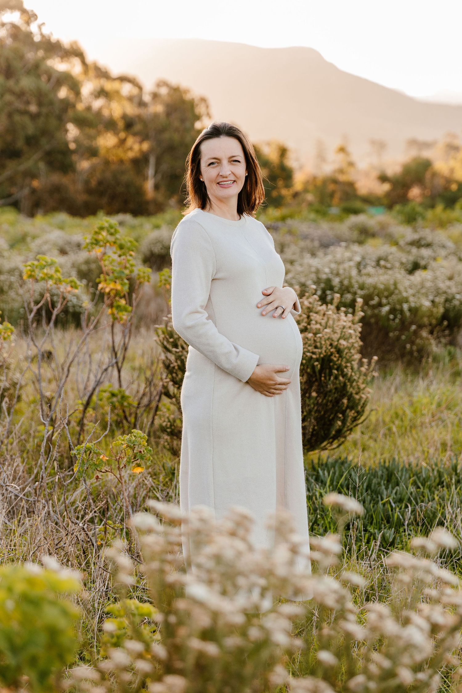 Pregnant woman in white dress cradling baby bump outdoors in golden hour light with mountains behind.