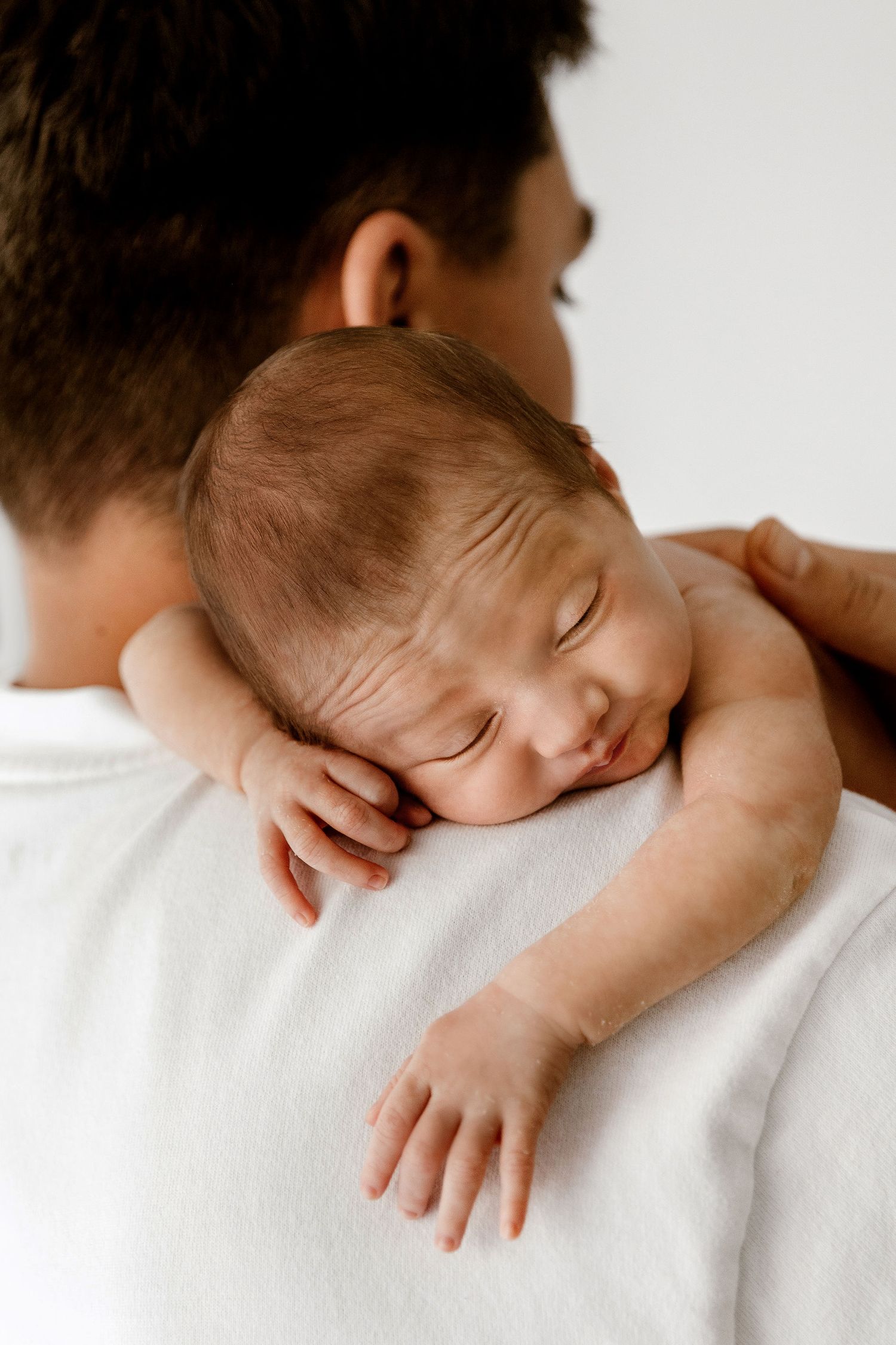 Peaceful newborn baby sleeping on father's shoulder, wearing white shirt, in a tender portrait photo.