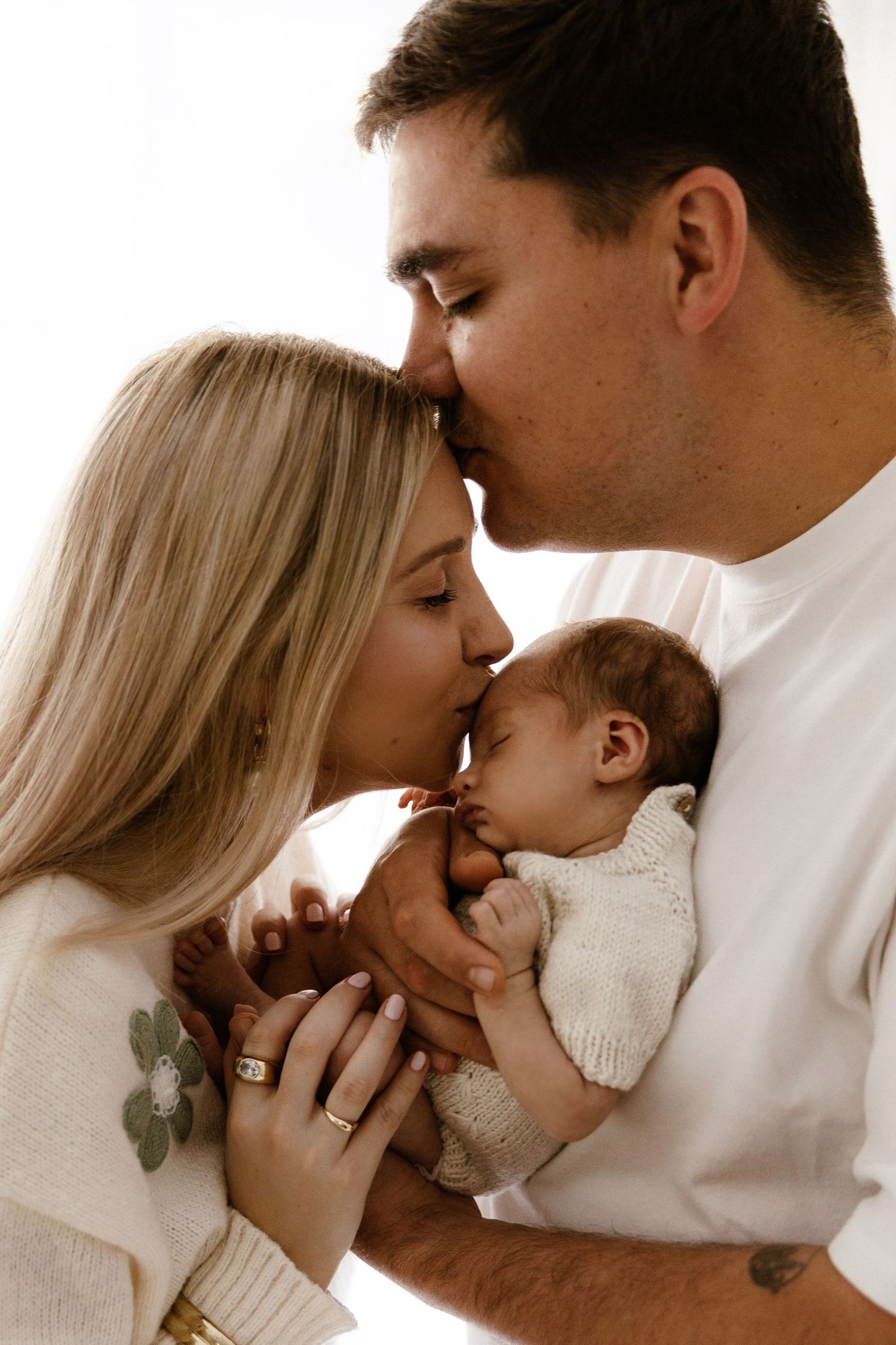 A couple in cream outfits share a tender kiss over their sleeping newborn baby in a bright, airy setting.
