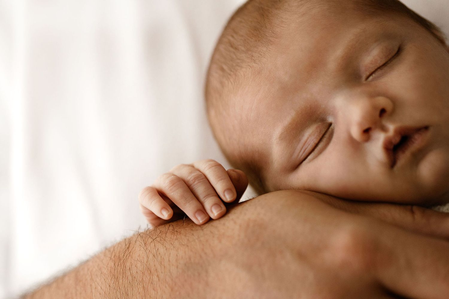 Sleeping newborn baby rests peacefully on parents arm in soft close-up photo.