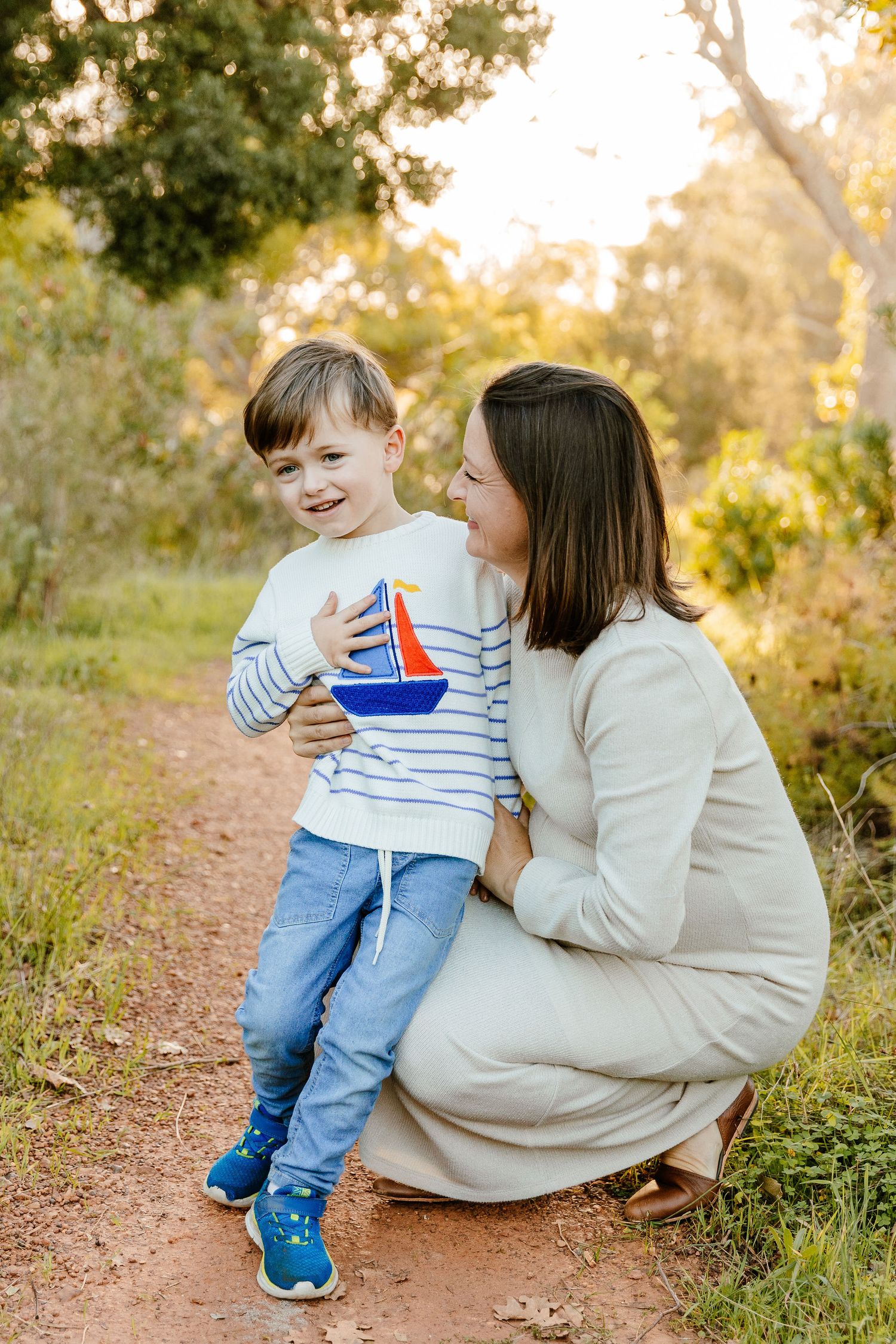 Mother crouching beside her young son holding a toy sailboat on a sunlit nature trail outdoors.