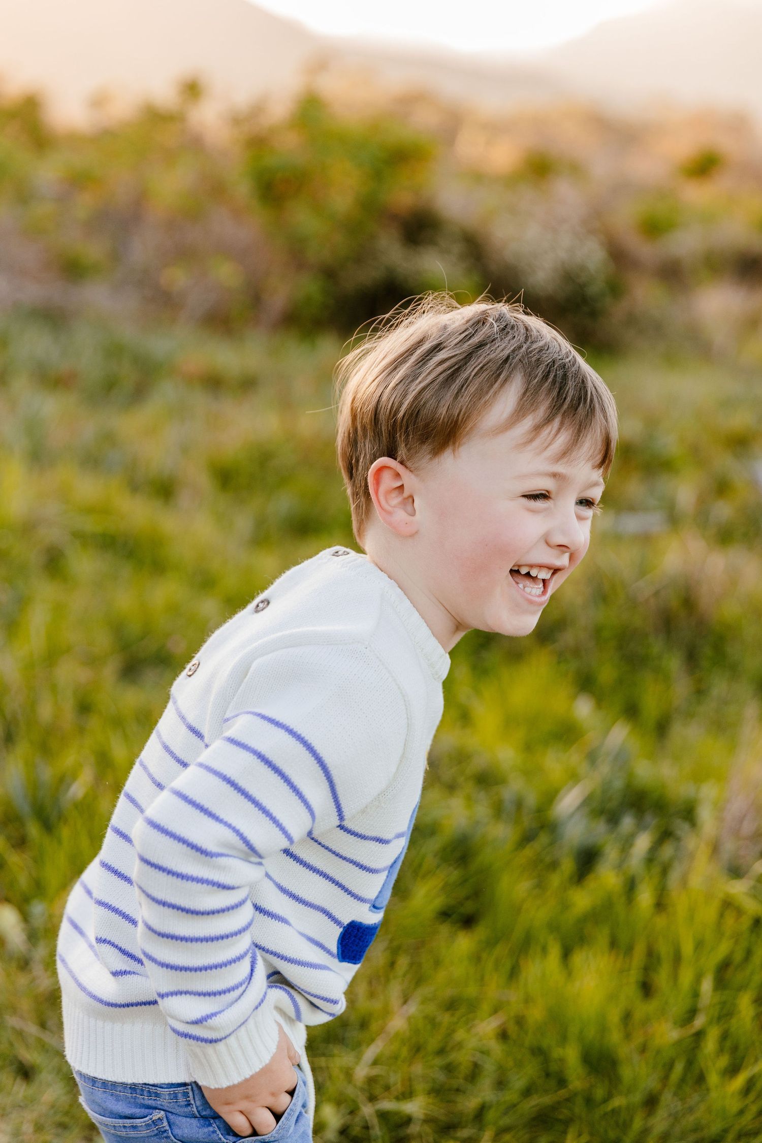 Cheerful boy in blue striped white sweater laughing while standing in a grassy meadow.
