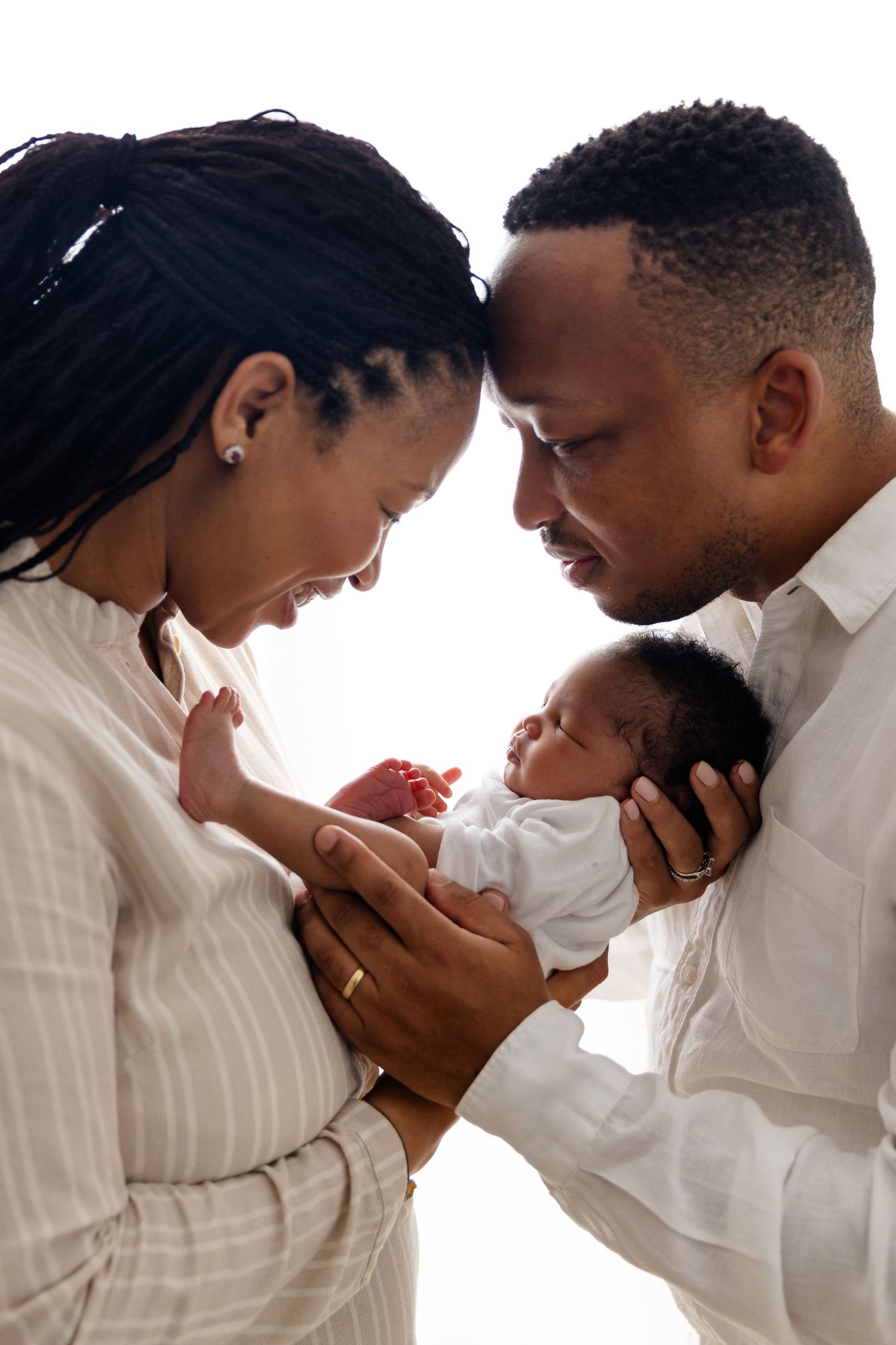 A couple tenderly holds their newborn baby together, leaning in close with loving smiles on a bright background.