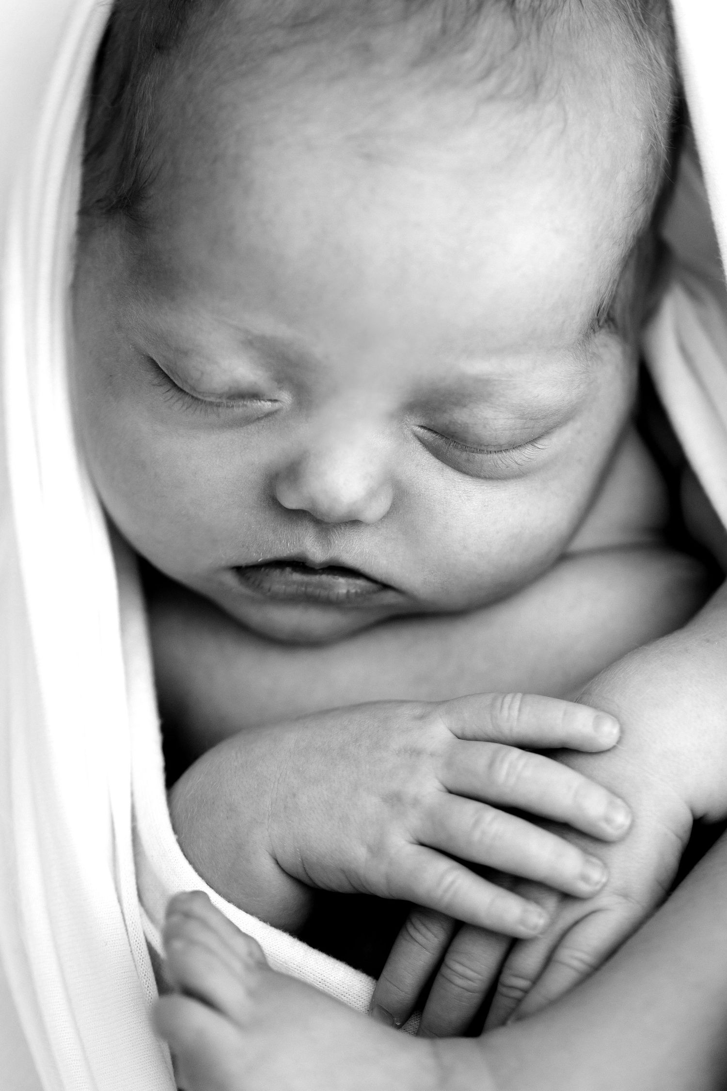 Black and white photo of a peaceful sleeping newborn baby wrapped in a soft white blanket with tiny hands visible.