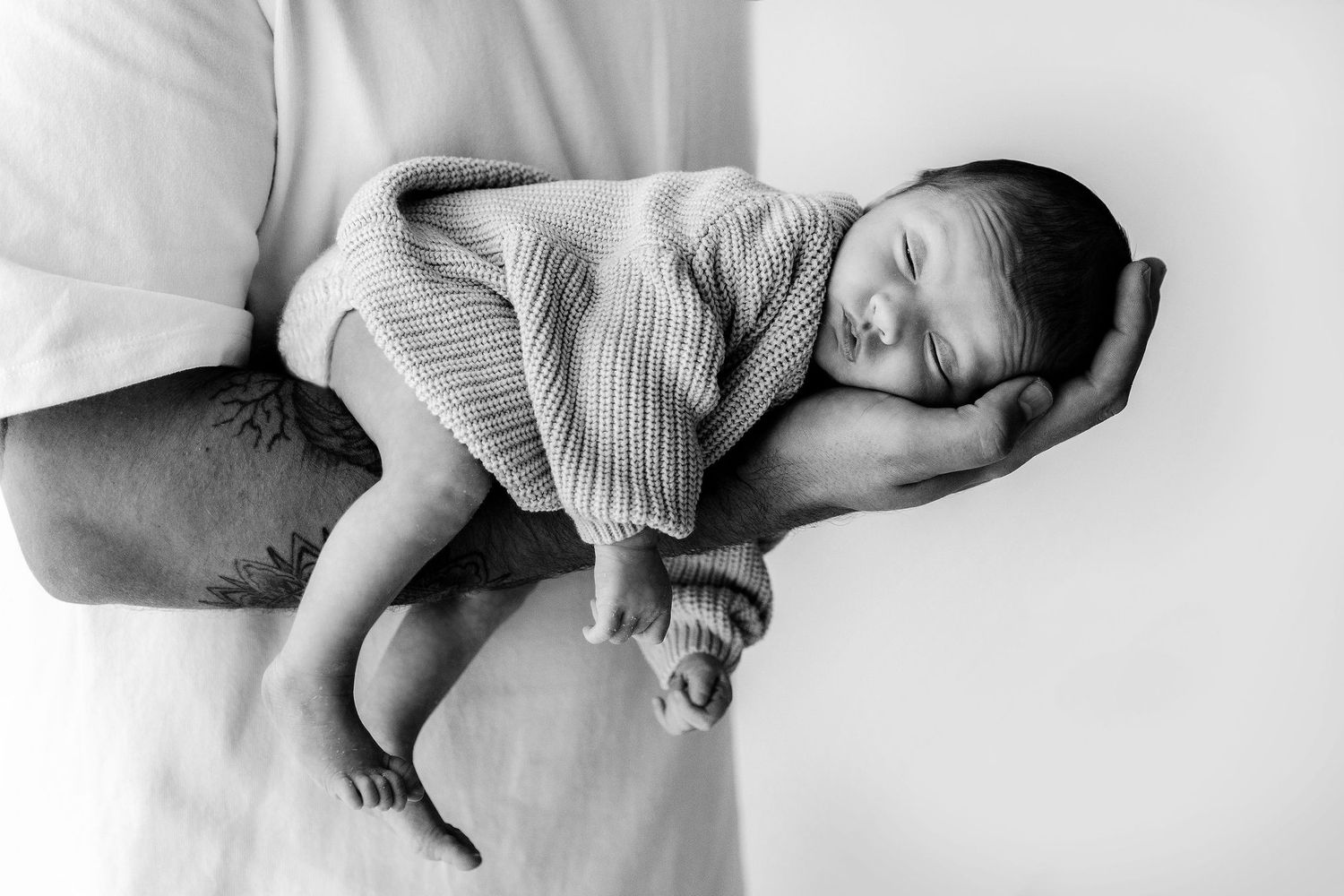 Black and white close-up of a sleeping newborn baby cradled in fathers tattooed hands.