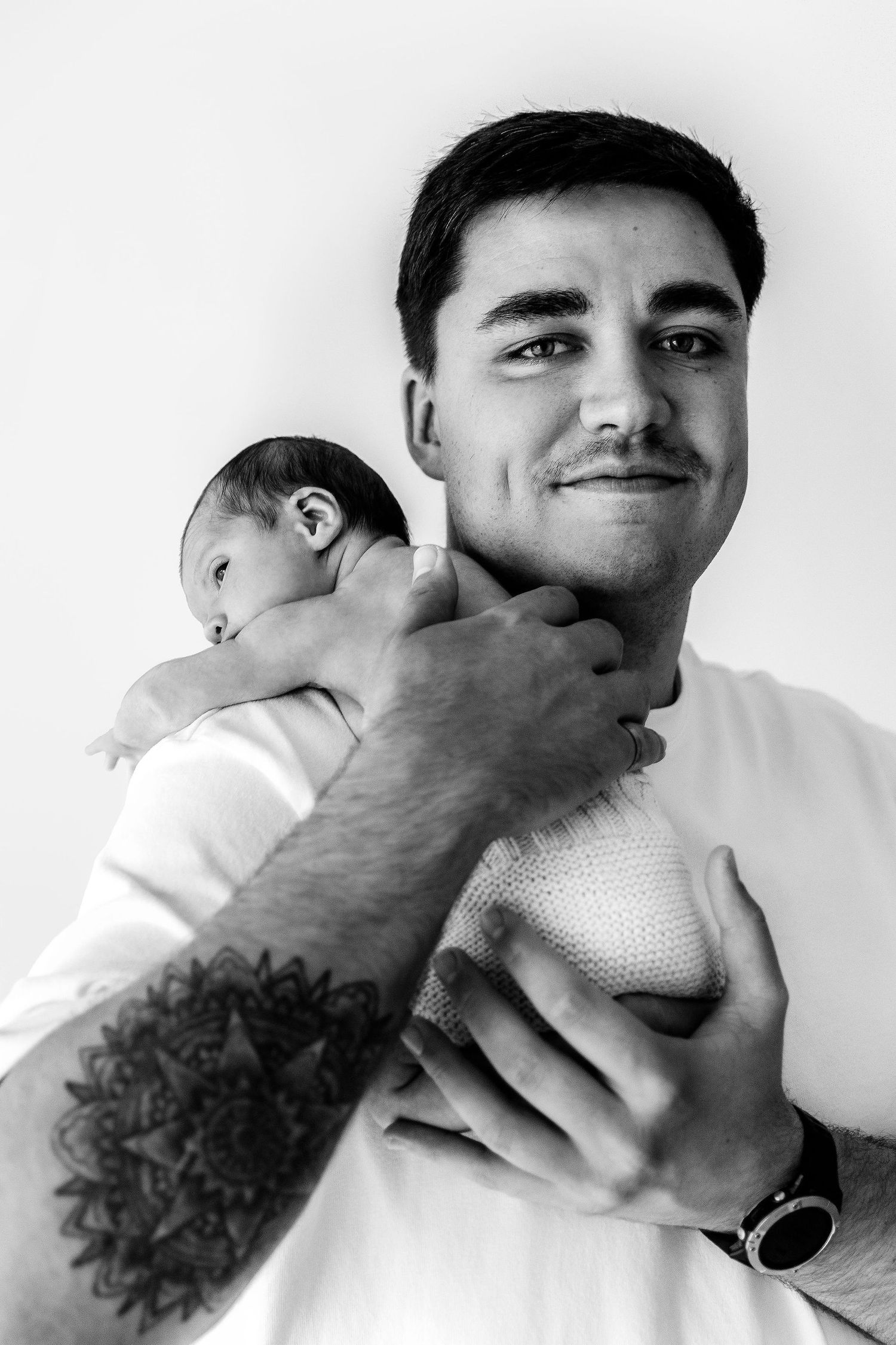 Black and white portrait of tattooed father holding newborn baby on his shoulder.
