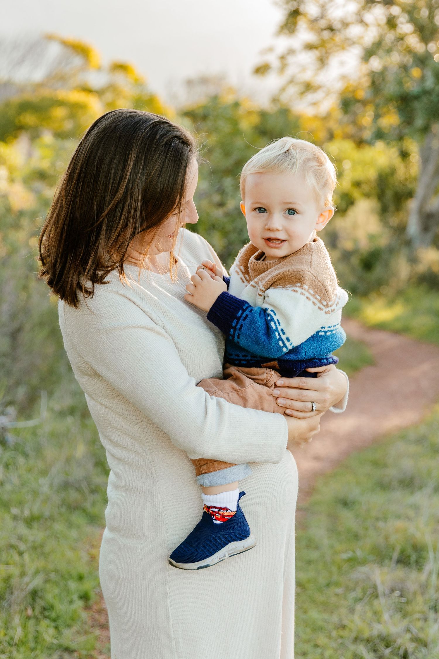 Mother in white dress holding her blonde toddler boy outdoors on a nature trail at sunset.