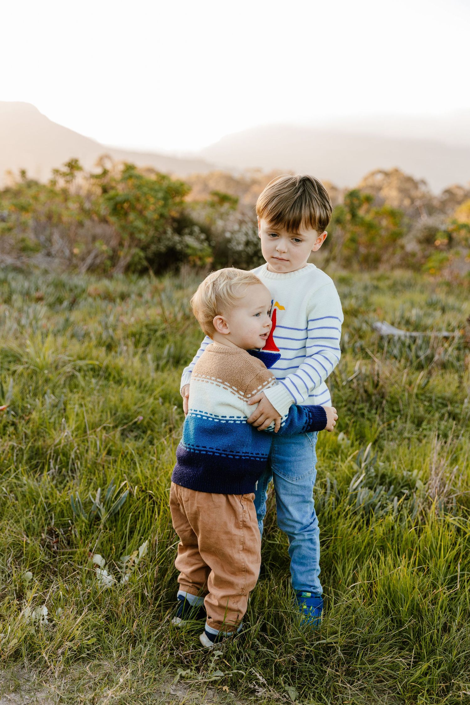 Two young brothers hugging in a grassy field with mountains and golden sunset light behind them.