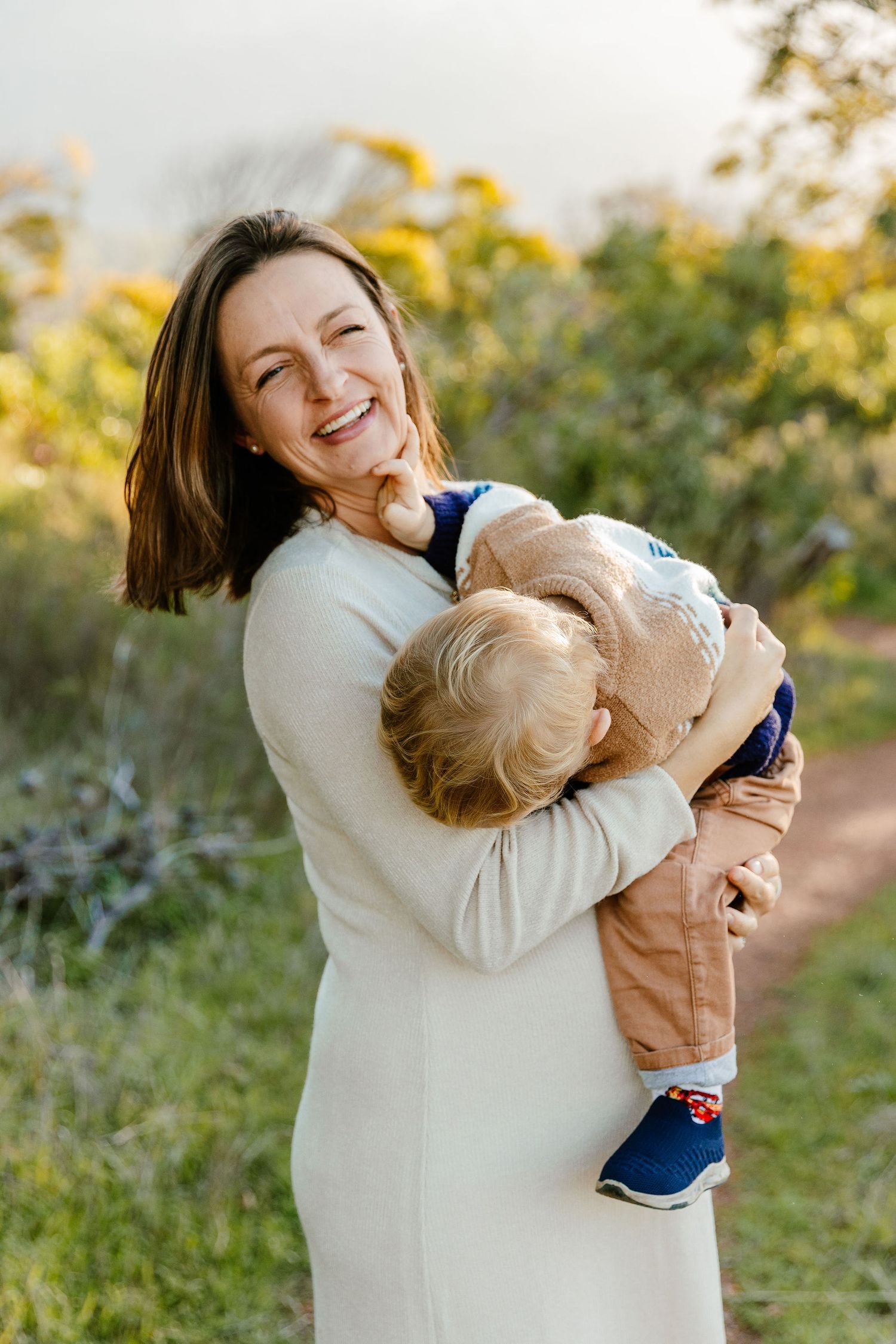 Joyful mother in white dress laughing while hugging her toddler boy outdoors in a green park setting.