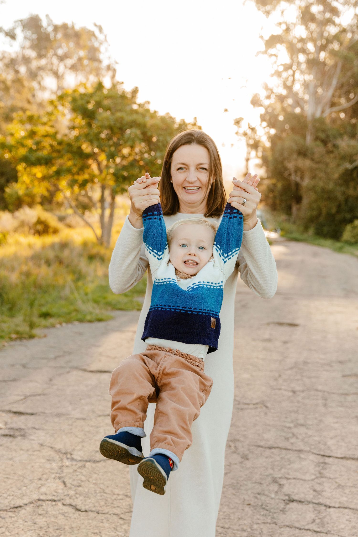 A smiling mother lifts her happy toddler boy outdoors on a sunny path surrounded by green trees.