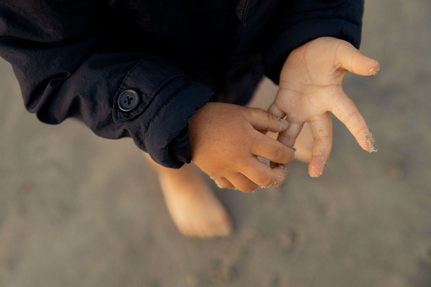 Close-up of a child's hands held together, wearing a dark blue jacket, standing barefoot on sandy ground.