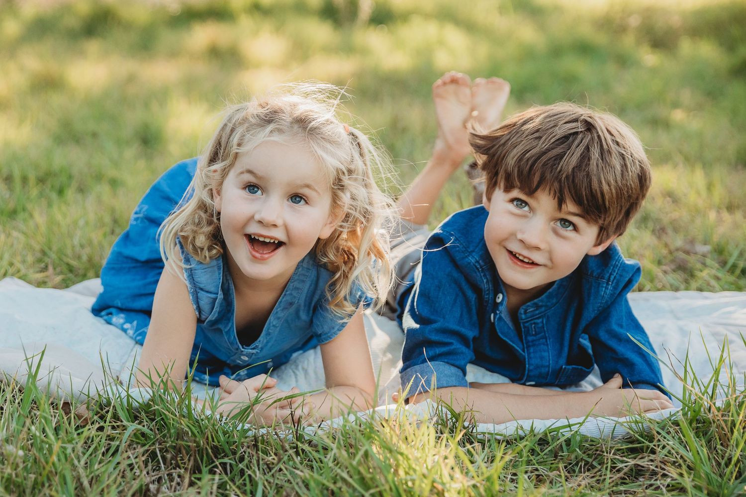 Two young siblings, a curly-haired blonde girl and a brown-haired boy, lying on a blanket in grass.