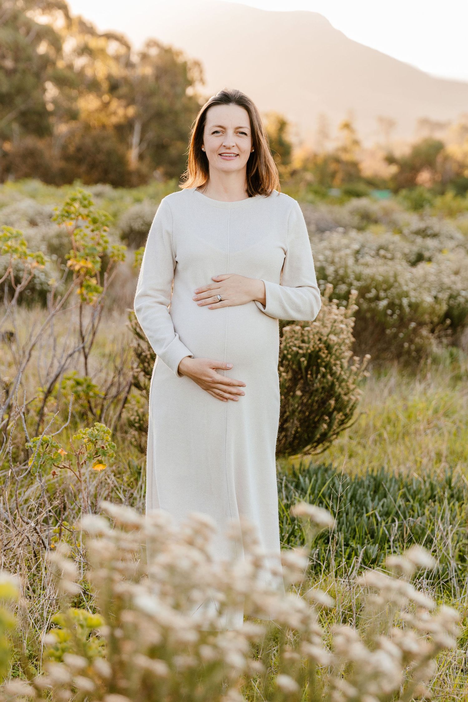 Smiling pregnant woman in white dress cradling baby bump in golden hour field with mountains.