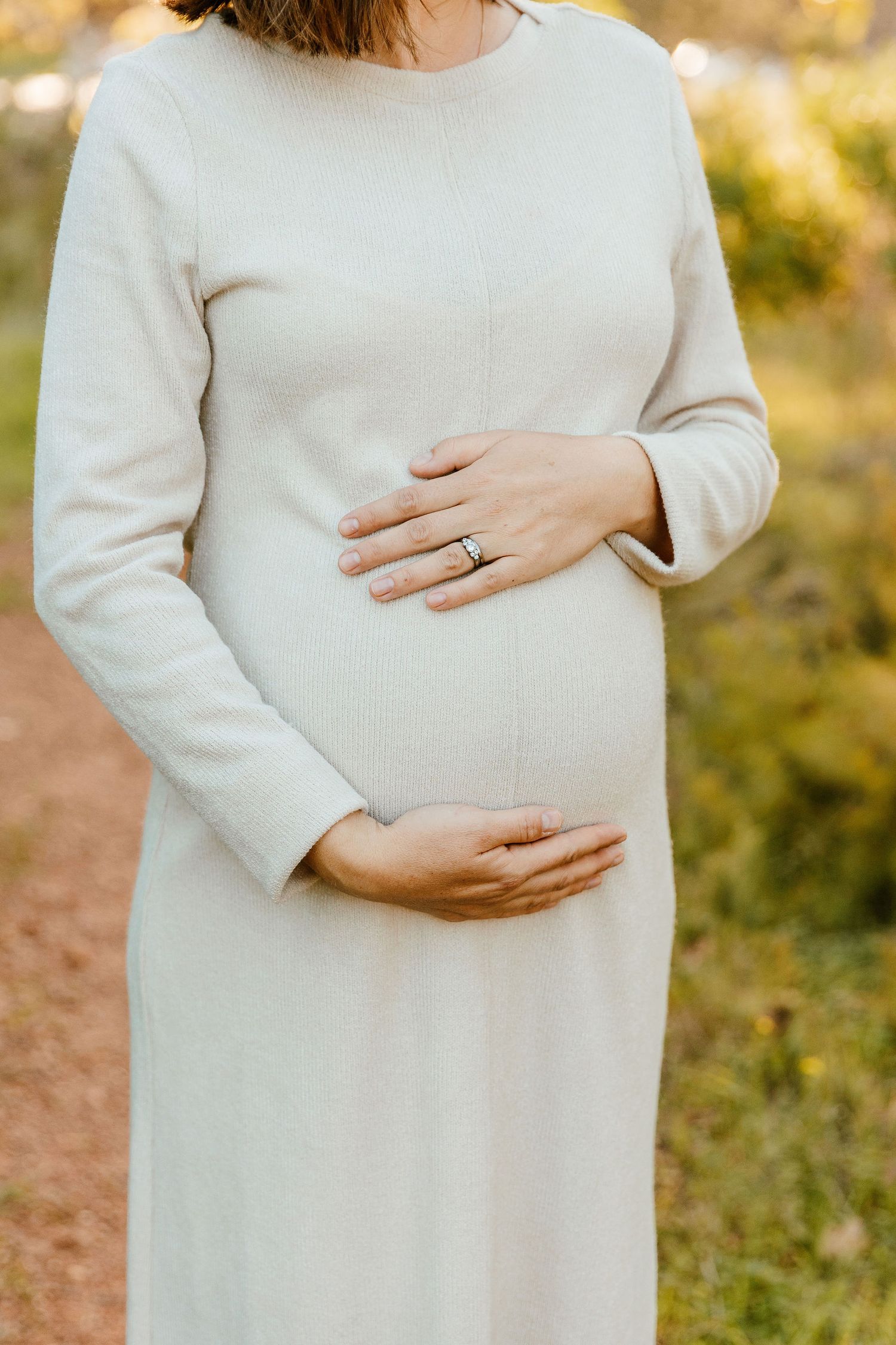 Pregnant woman in white dress cradling baby bump outdoors, wearing wedding ring, with green foliage background.