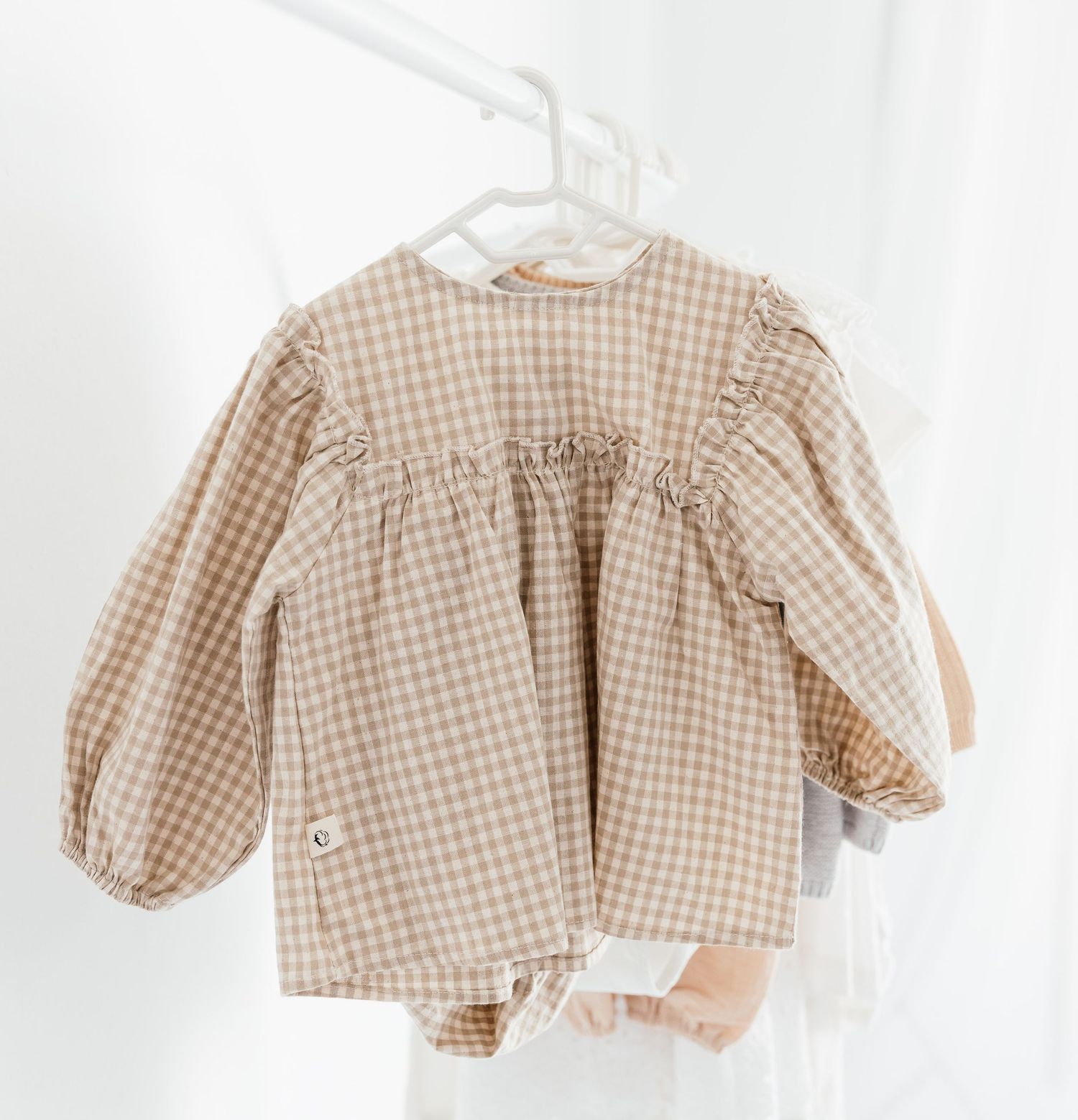 Beige gingham check baby blouse with ruffled yoke and balloon sleeves hanging on a white hanger.