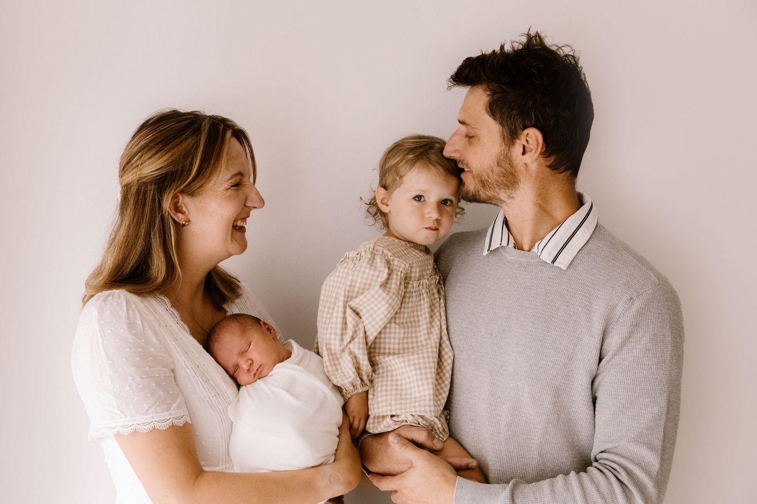 Happy family portrait with mother holding newborn baby and father carrying toddler girl against white background.