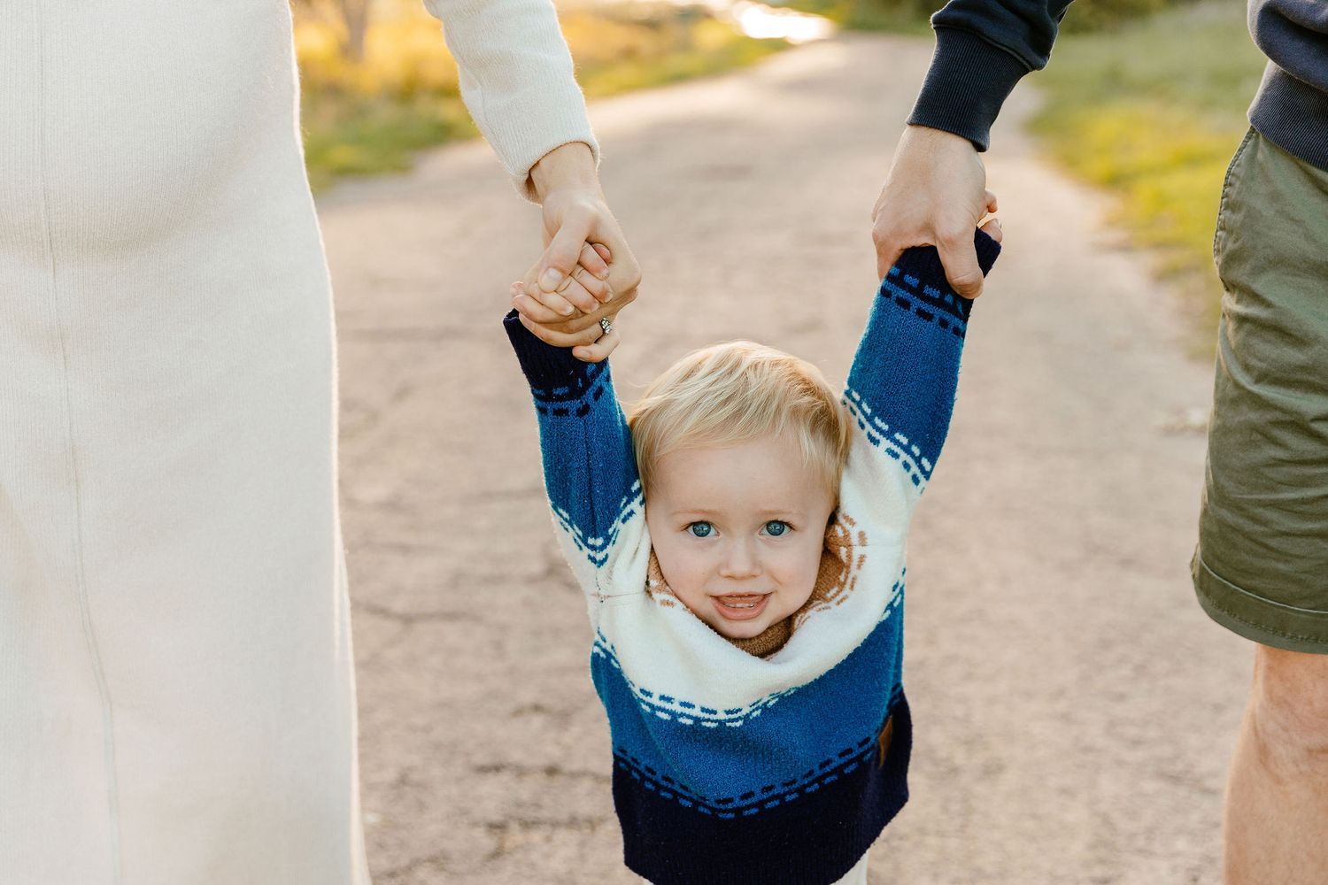 Toddler in blue sweater held by parents hands on a golden-lit path.