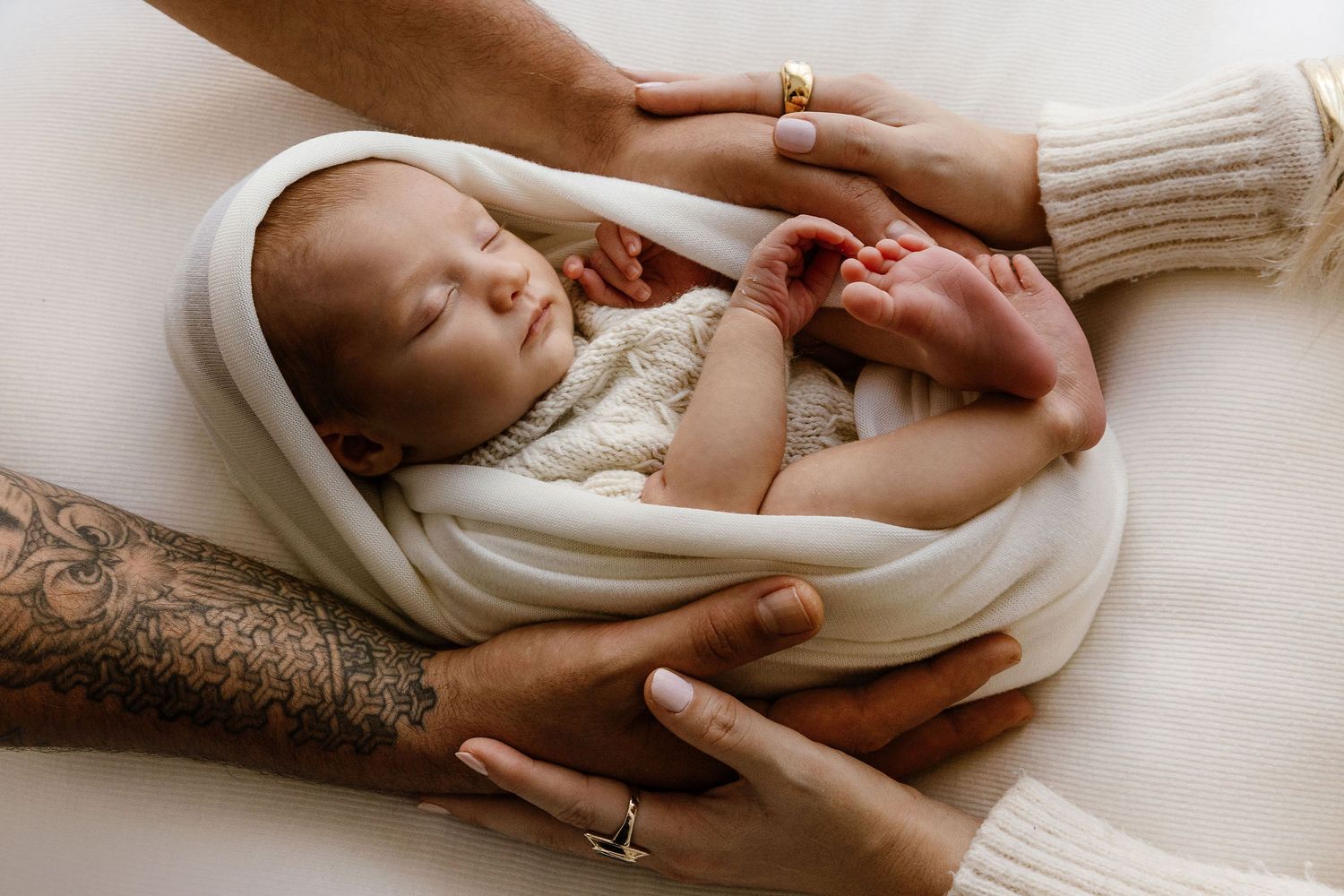 Parents' hands gently holding swaddled sleeping newborn baby, father with tattoo visible.