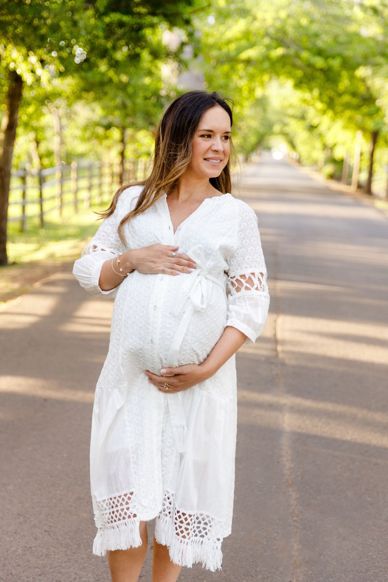 Smiling pregnant woman in white lace dress cradling baby bump outdoors on tree-lined path.
