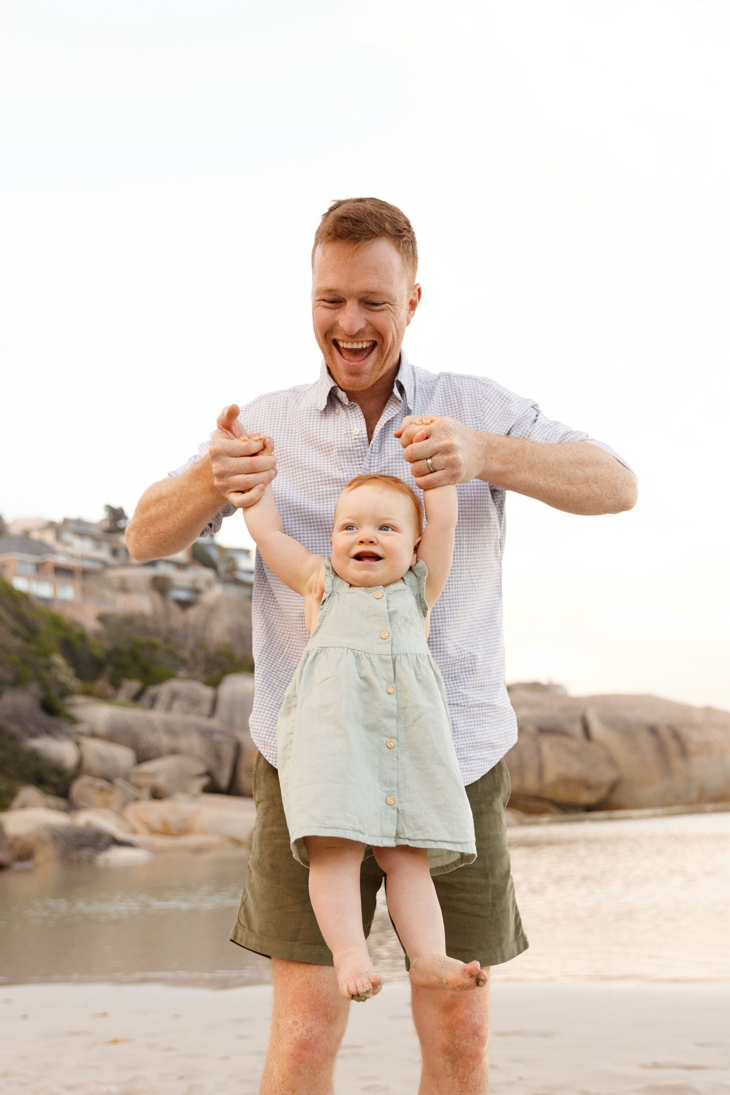 Laughing father lifting happy red-haired baby girl on sandy beach.