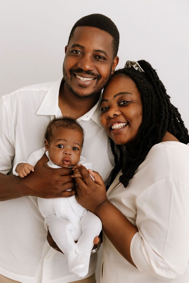 Happy Black family portrait with smiling parents holding their newborn baby, all dressed in white.