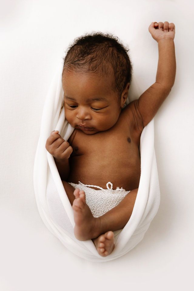 Peaceful newborn baby wrapped in white cloth with one arm raised, lying on white background.