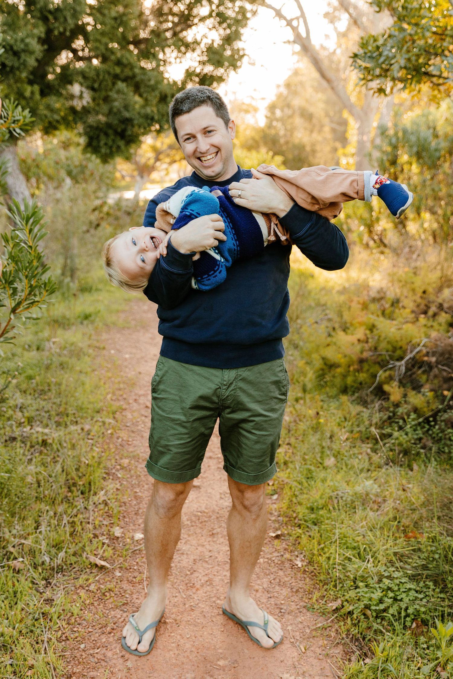 Laughing father playfully holds toddler upside down on a sunlit nature trail, wearing navy sweatshirt and green shorts.