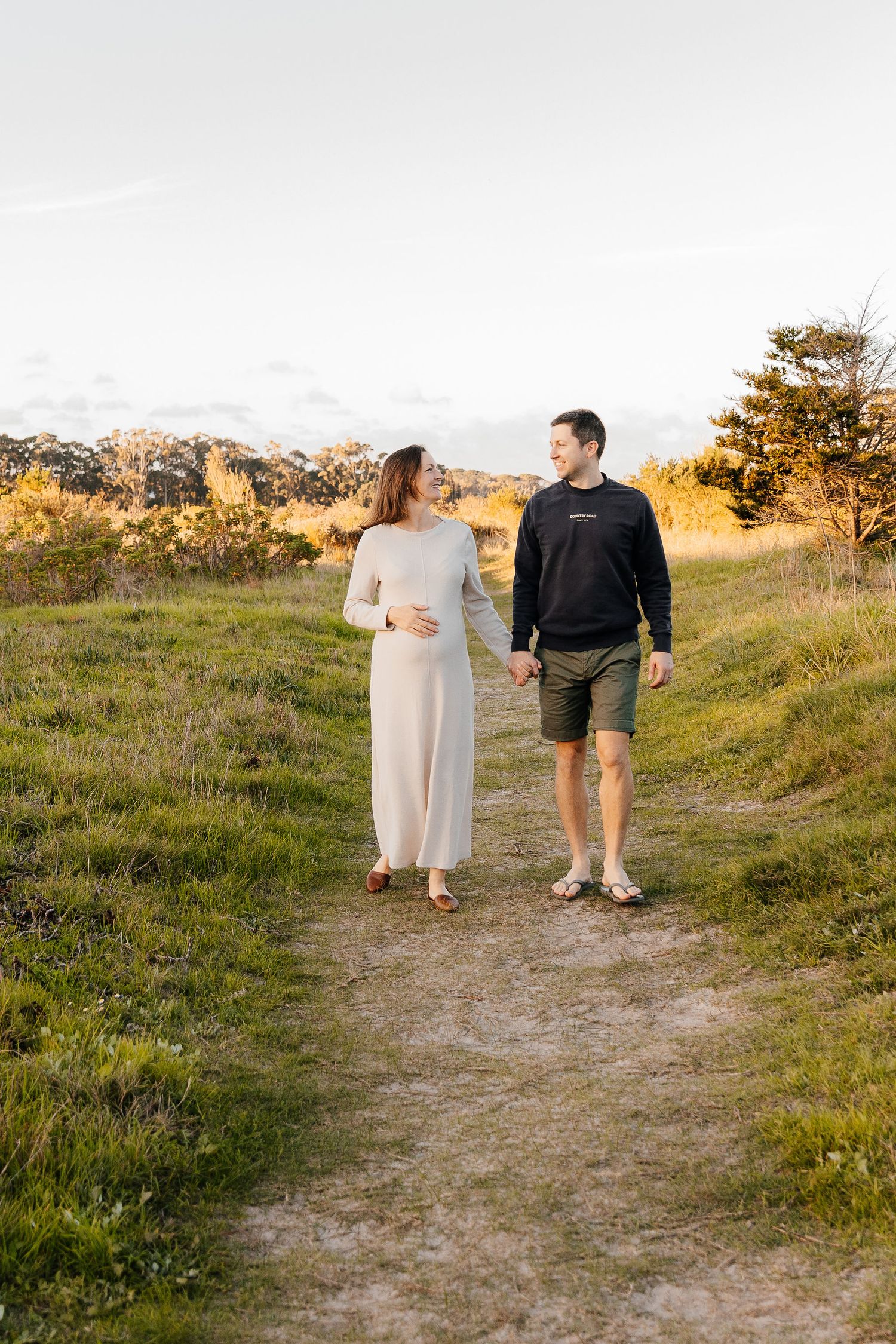 Expecting couple walking hand in hand on a dirt path through a green field at golden hour.