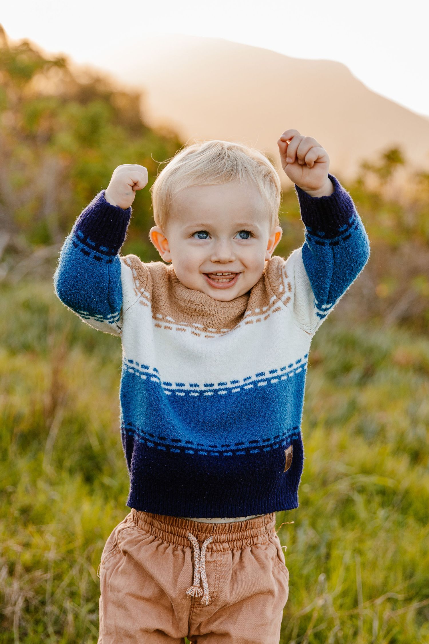 Excited toddler boy raising both fists in the air wearing a striped knit sweater in a green meadow.