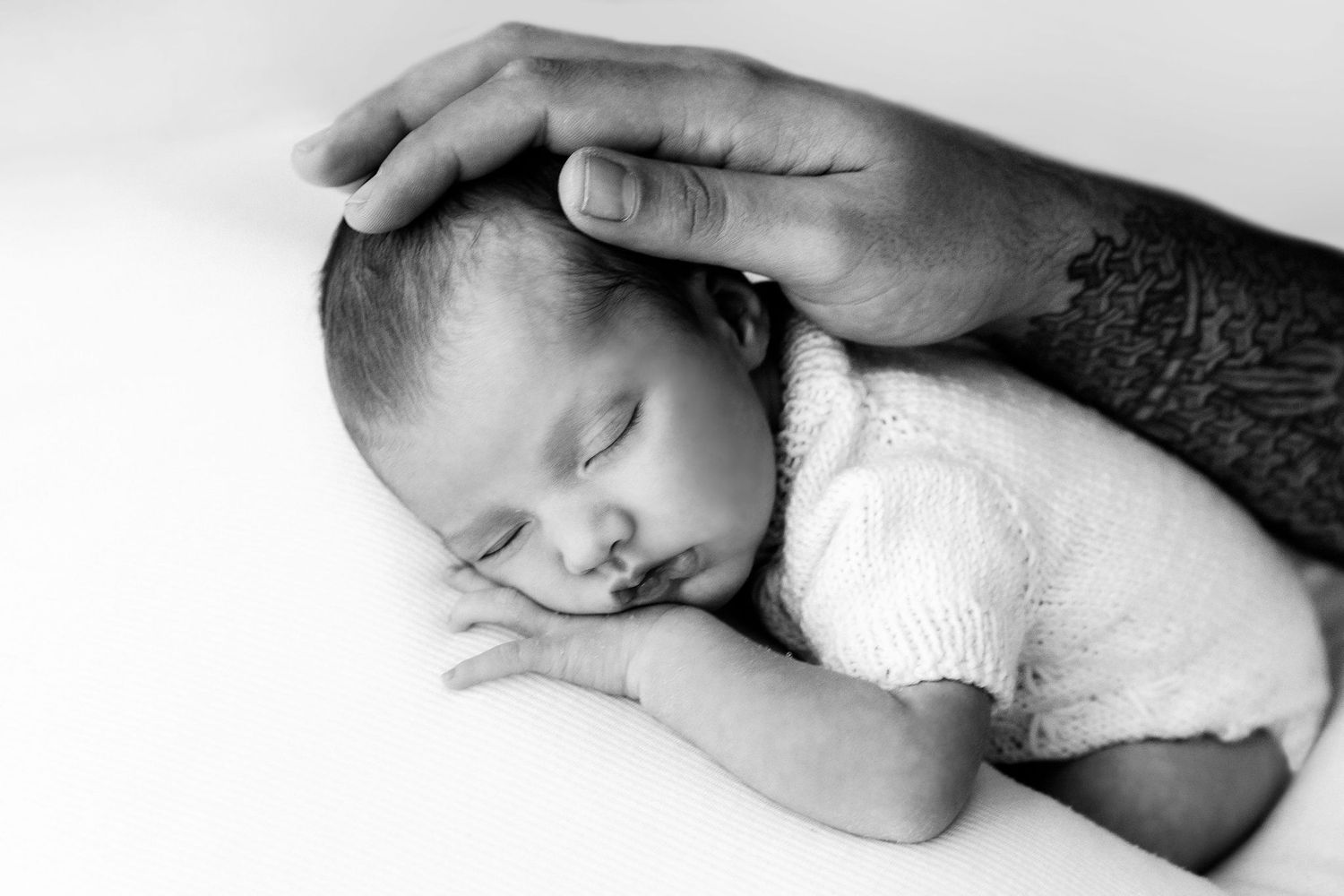 Sleeping newborn baby in knit romper with fathers tattooed hand on head.