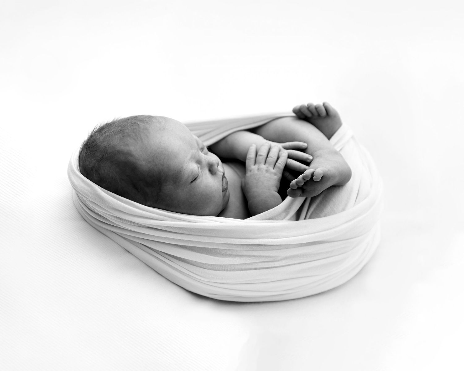 Black and white newborn photo of a sleeping baby swaddled in white wrap, curled in fetal position.