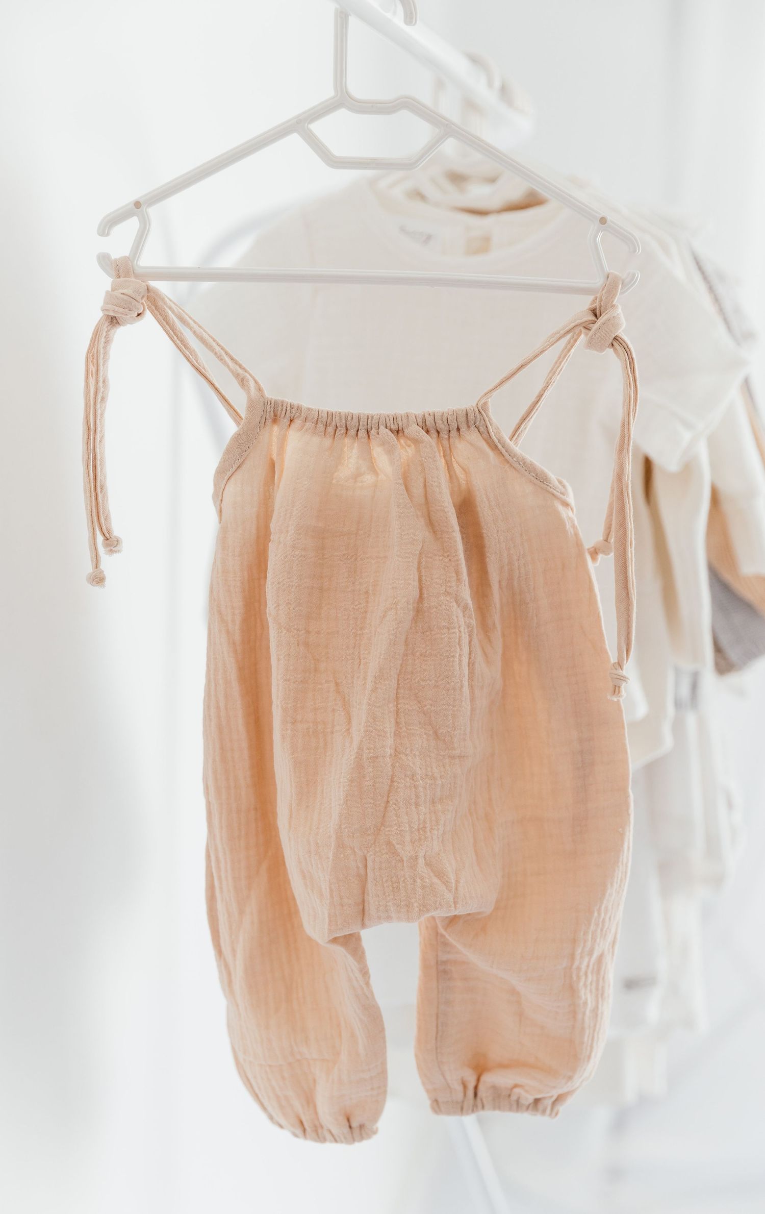 Peach muslin baby romper with tie straps hanging on a white hanger, soft neutral tones, minimalist style.