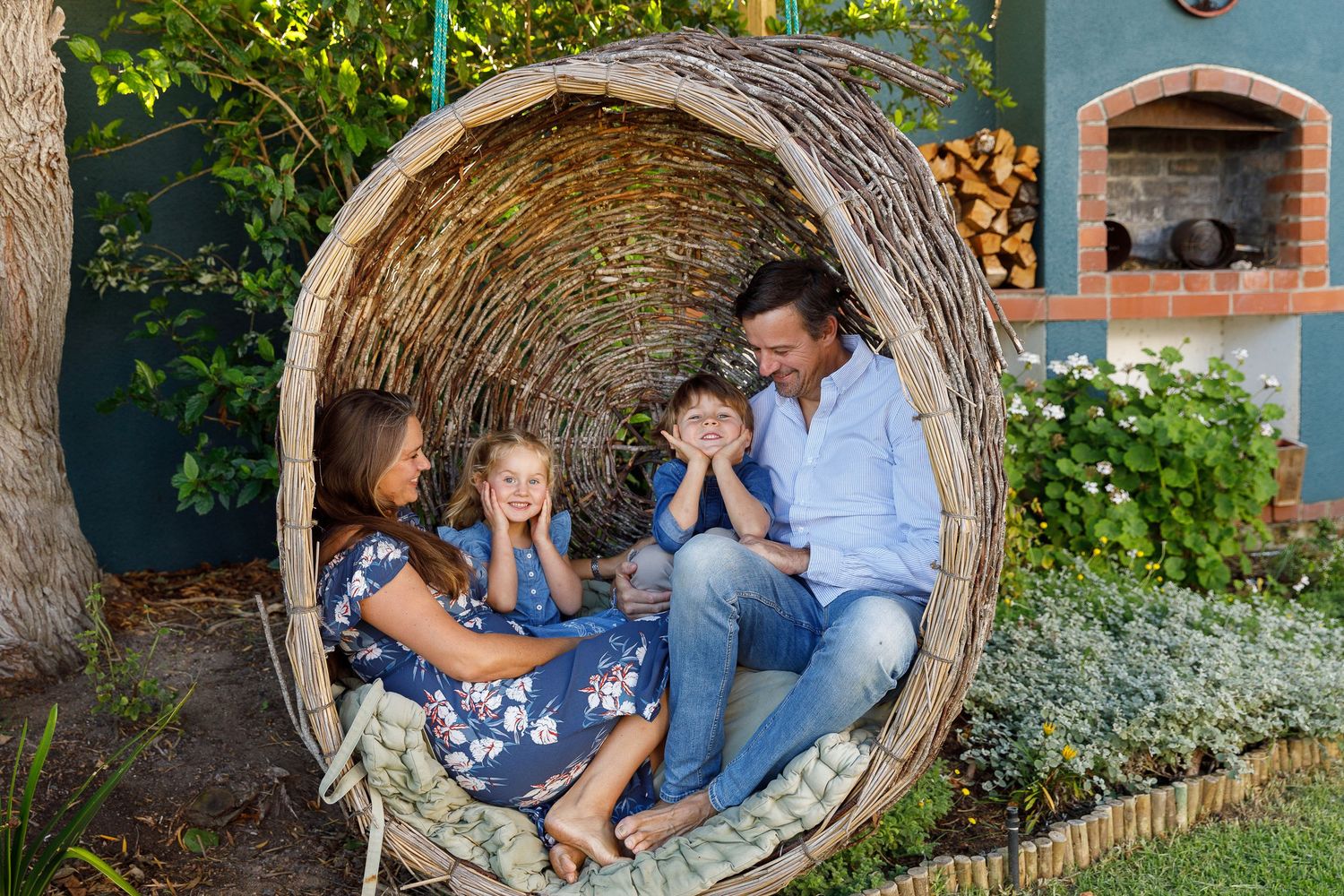 Family of four laughing together inside a large woven nest garden feature with pizza oven in background.