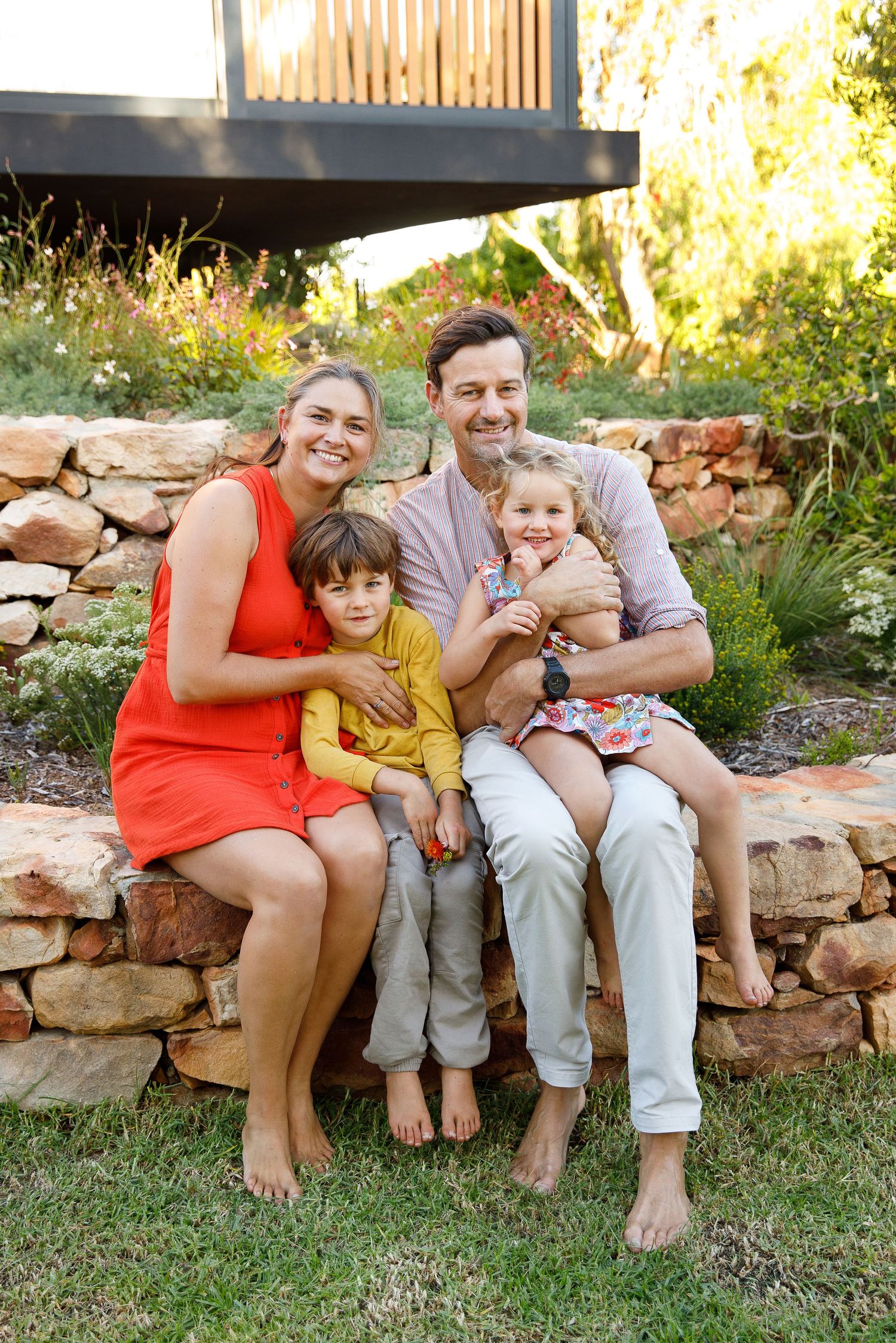 Happy family of four sitting barefoot on a stone garden wall surrounded by lush plants.