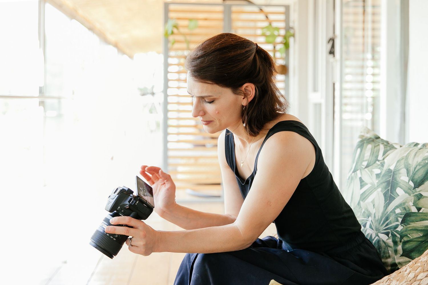 Woman photographer reviewing photos on DSLR camera screen while sitting indoors in bright natural light.