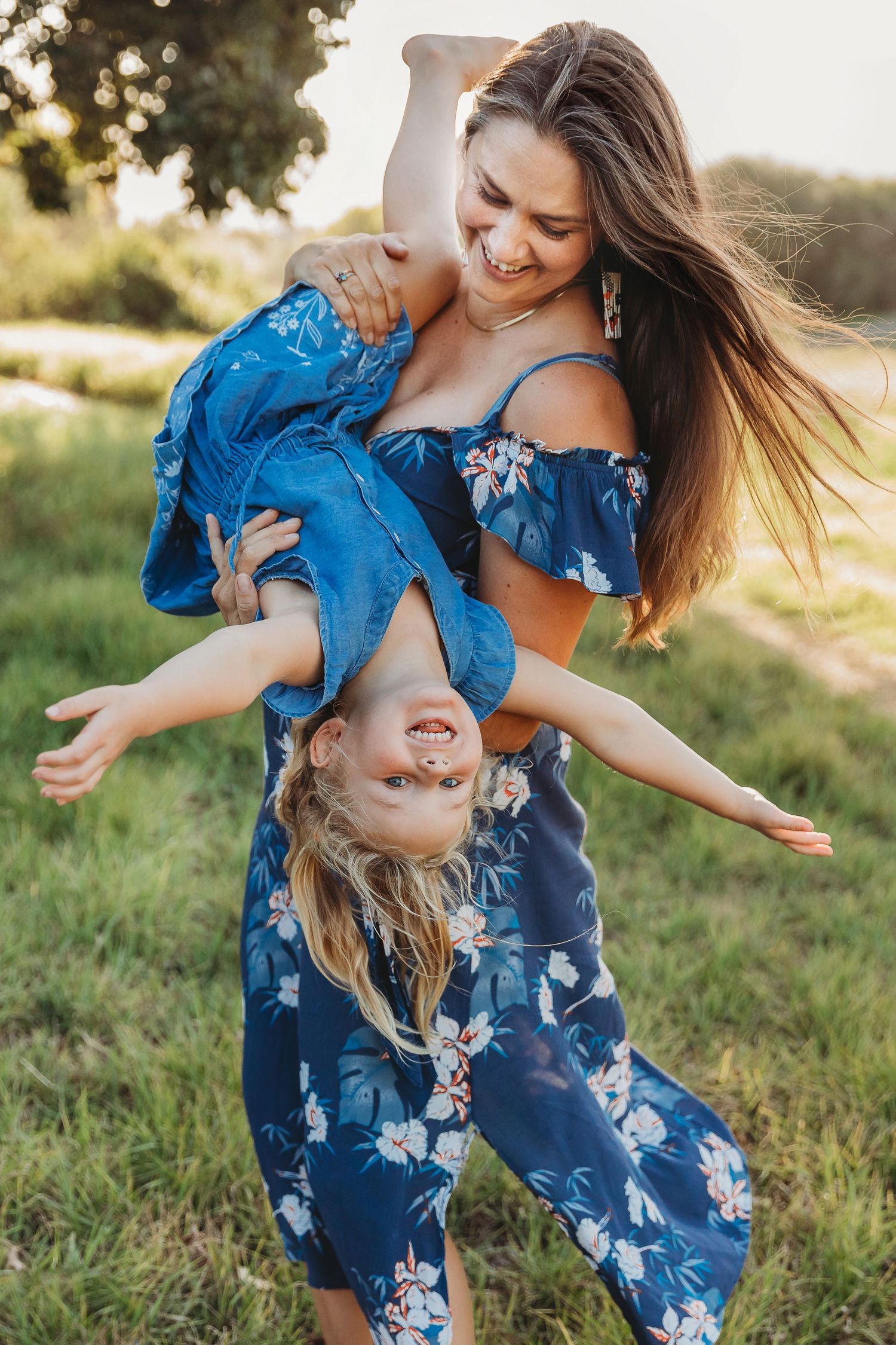 A smiling mother in a blue floral dress playfully holds her laughing daughter upside down outdoors.