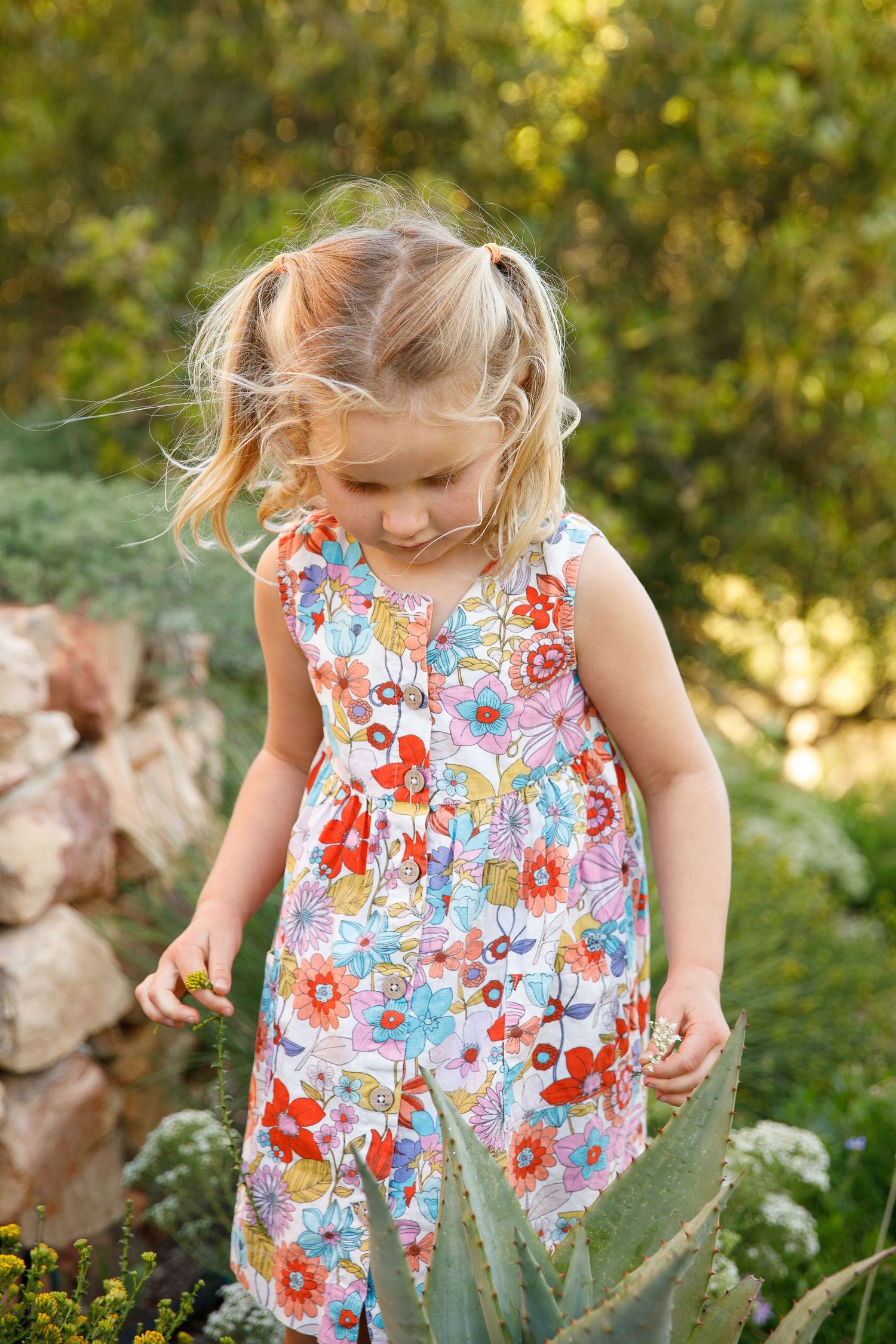 Young blonde girl in colorful floral dress exploring a lush garden, looking down at plants.